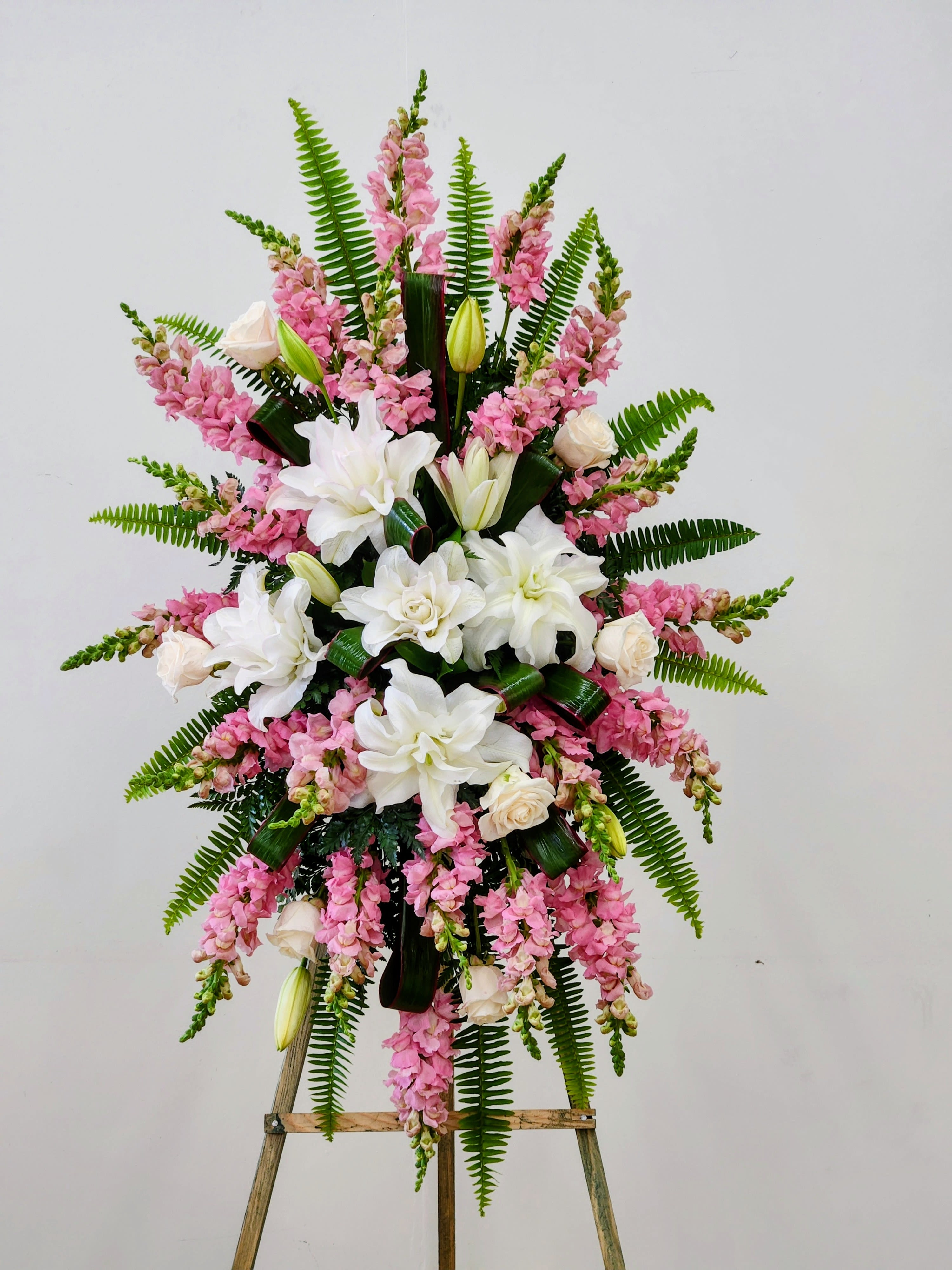 Pink Remembrance Spray - A tall, graceful spray of white lilies and pale cream roses framed by cascading pink snapdragons-this standing arrangement is designed as a sympathetic tribute for memorials and funeral services. The Pink Remembrance Spray reads as quietly dignified and ceremonial, the soft pinks and whites conveying tenderness and respect for the person you're honoring.  Suitable for a funeral, memorial service, or graveside tribute, this arrangement is the kind of floral expression that helps a grieving family feel seen and supported. Delivered on a sturdy wooden easel and arranged for presentation, it arrives ready to display; please let us know any service details or personalization requests when ordering so we can ensure timely delivery.