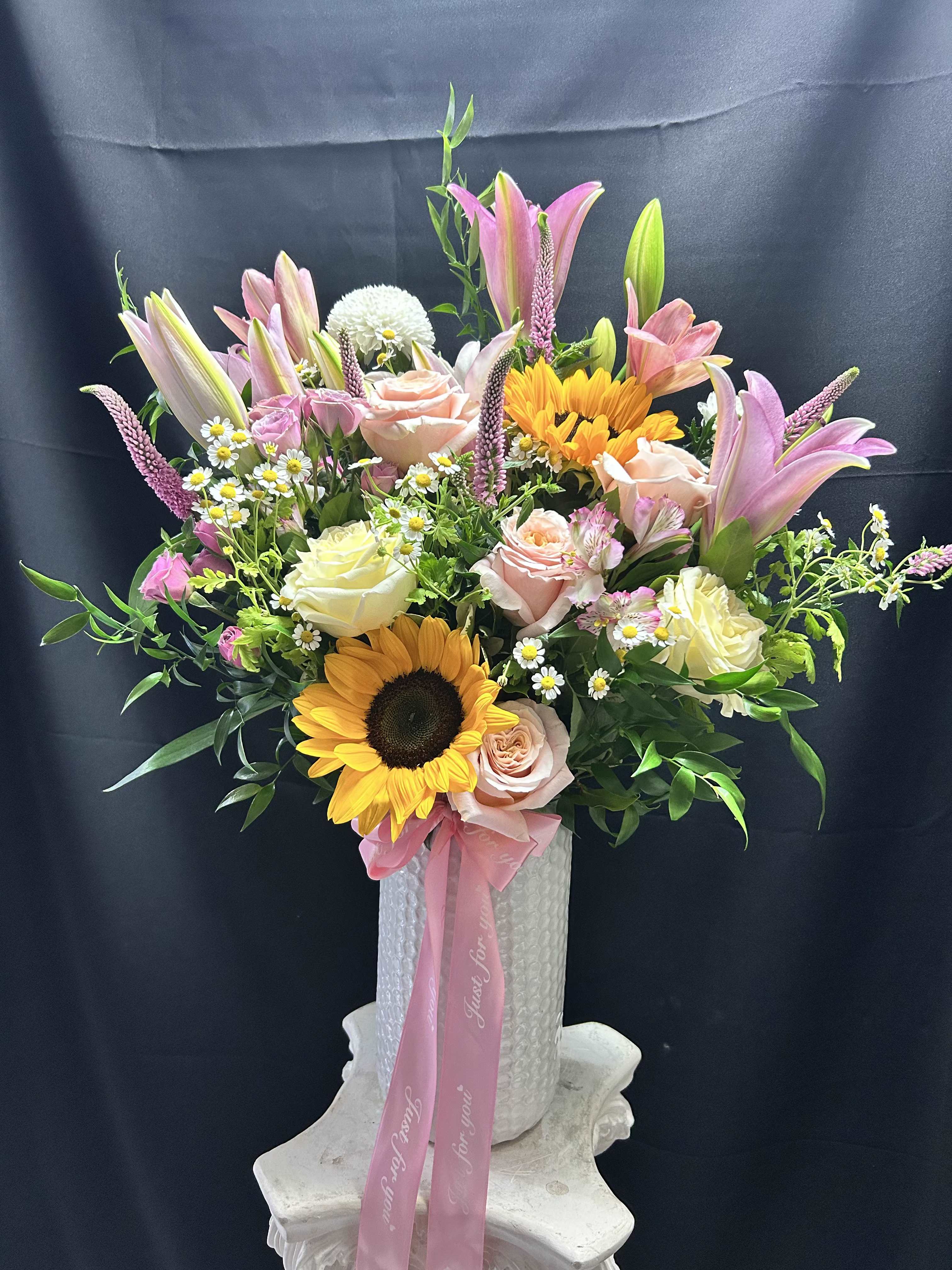 Spring is here  - Sun-kissed sunflowers and tall pink lilies give this upright mixed flower arrangement an uplifting first impression-perfect as a birthday bouquet, a Mother's Day gift, or an anniversary surprise. The combination of bright yellow focal blooms with soft blush roses, creamy alstroemeria, snapdragons and small white daisy-like fillers creates a lively yet romantic look that reads as both celebratory and heartfelt for love and romance, thank yous, or just-because moments.  Arranged full and tall in a white textured vase and finished with a pink ribbon, the composition adds height and movement through open blooms and vertical spikes for a naturally airy silhouette. The palette of pink, yellow, white and soft green offers cheerful contrast and gentle texture, making it equally right for housewarming, congratulations, or to brighten someone's day.  This gift-ready presentation arrives polished on a decorative pedestal-ready vase, so the person you're thinking of will feel genuinely celebrated the moment it's set out. Order as shown for a large, abundant arrangement that's easy to care for-fresh water in the vase and a cool spot away from direct heat will help blooms last.