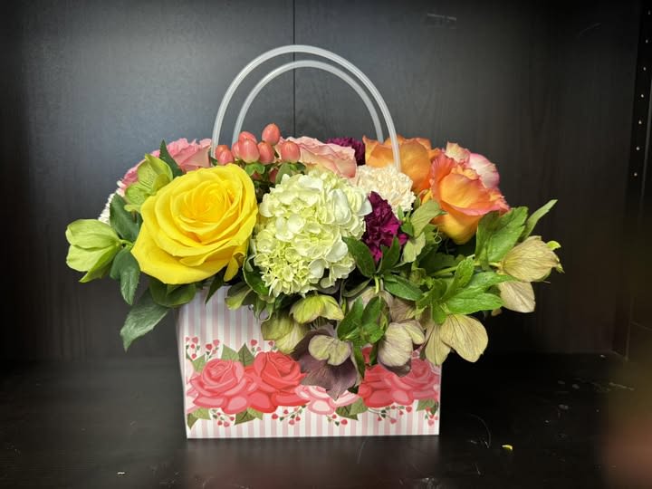 Mom called - she loves this! - Mom called and she wanted us to tell you that she loves this!  The arrangement is presented in a decorative handled gift bag that resembles a purse, ready to hand over, and reads bright and celebratory so the recipient feels seen and delighted the moment they see it.  Inside the bag you'll find a dense, garden-like mix of roses, hydrangeas, carnations and other seasonal blooms in yellow, pink, white, orange and deep magenta. Pale hydrangea and clustered carnations add soft volume through the center while the roses and brighter accents create focal points and texture. Sized as a hand-held gift bag arrangement, it travels well and makes an immediate, joyful impression-no vase required.