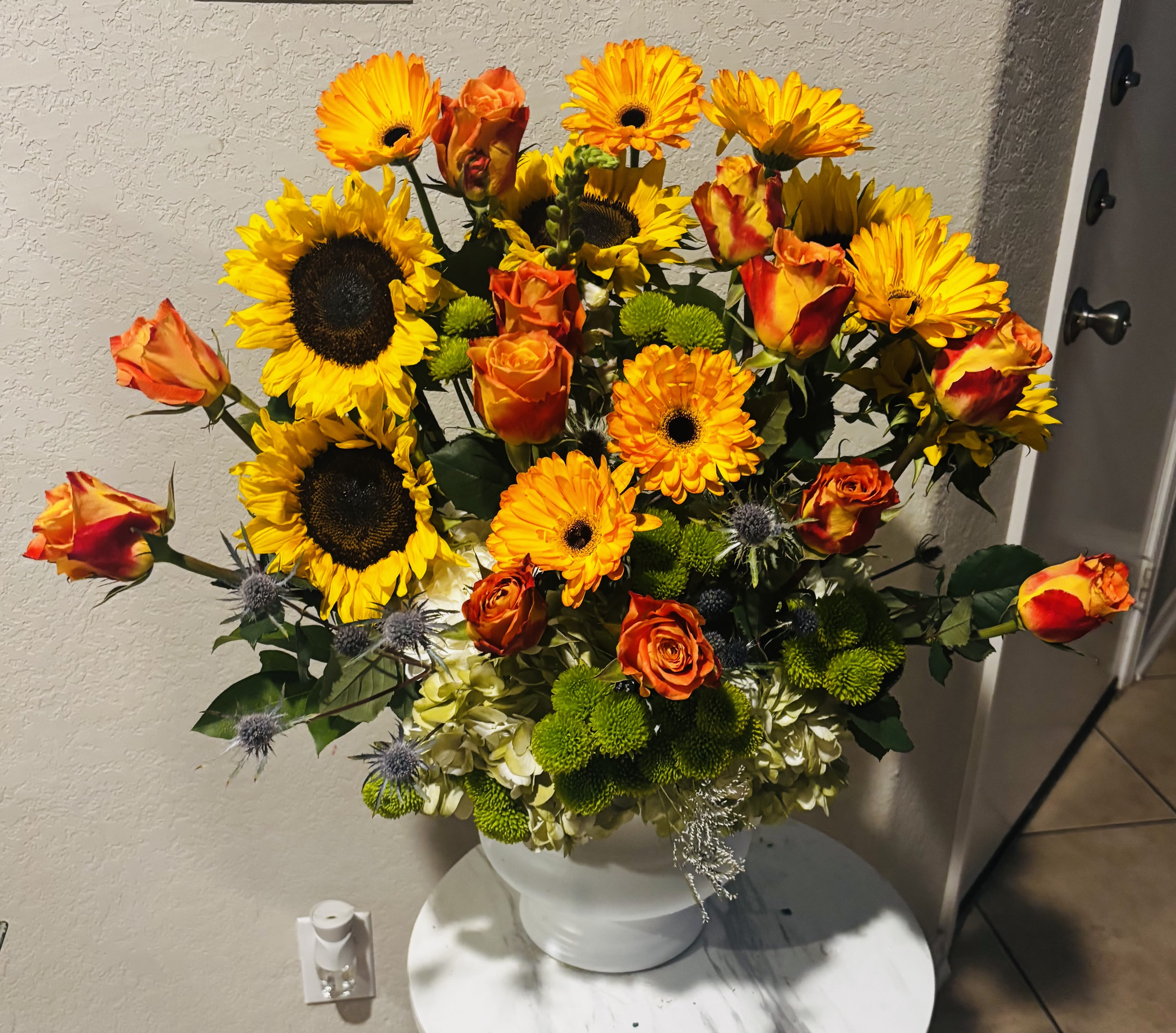 Sunflower &amp; Orange Rose Luxury Arrangement - A vibrant sunflower flower arrangement in San Antonio featuring fresh sunflowers, orange roses, gerbera daisies, green button mums, hydrangeas, thistle, and premium seasonal greenery beautifully designed in an elegant white urn vase. Handcrafted by Snyder Flowers San Antonio, this bold and cheerful floral arrangement is perfect for birthdays, celebrations, congratulations, get well wishes, fall decor, or meaningful gifts.   We proudly offer same-day flower delivery in San Antonio with fresh premium flowers and exceptional customer service.  Luxury floral designs created with high-quality blooms, bright seasonal colors, and elegant modern garden style.  Serving Stone Oak, Medical Center, Alamo Heights, Cibolo, Universal City, Live Oak, Helotes, Castle Hills, Shavano Park, and Downtown San Antonio.  Looking for a trusted florist in San Antonio, same day flower delivery, or sunflower arrangements near you? Snyder Flowers creates elegant handcrafted flower arrangements with fresh roses, sunflowers, hydrangeas, and premium blooms designed to impress for every occasion.