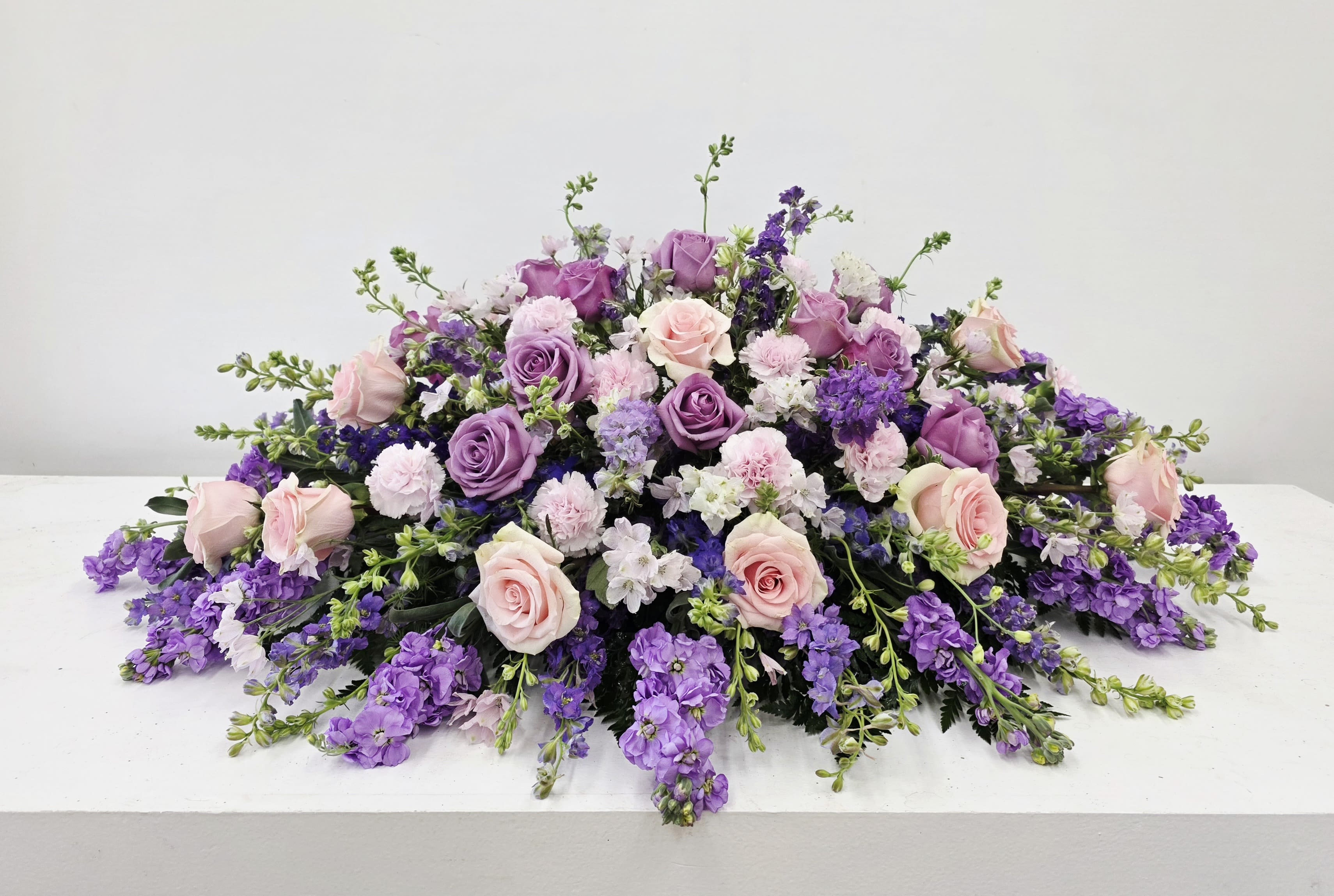 Sweet Memories Casket Spray - Soft lavender and blush come together in a low, broad floral design perfect for a sympathy arrangement. This bouquet-style spray places lavender and purple roses front and center, surrounded by pale carnations and clusters of tiny pale pink and lavender blooms that create a layered, garden-fresh silhouette - the kind of arrangement that makes the person you're honoring feel tenderly remembered.  