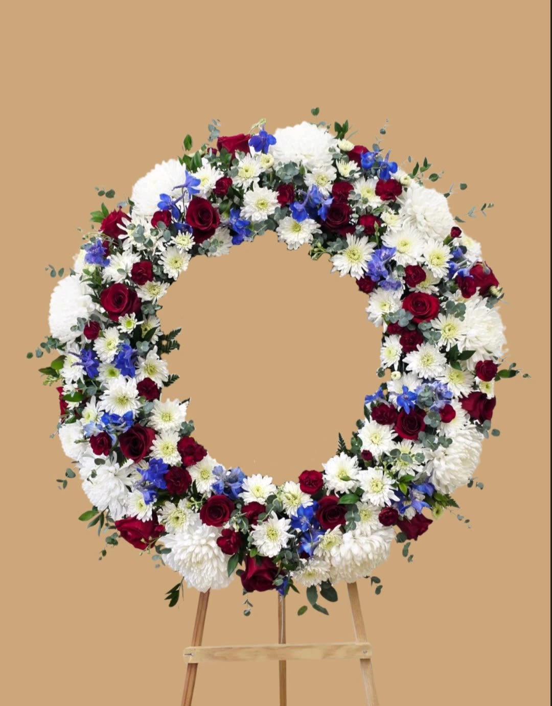 Honor in Service Wreath - A formal circular wreath of deep red roses, snowy white chrysanthemums and daisies, and bright blue delphinium-like accents arranged on a sturdy wooden easel-designed as a respectful floral tribute for memorials, veterans' services, or patriotic ceremonies. The red, white, and blue palette reads clearly from a distance, creating a solemn, polished impression that honors the person you're remembering.  