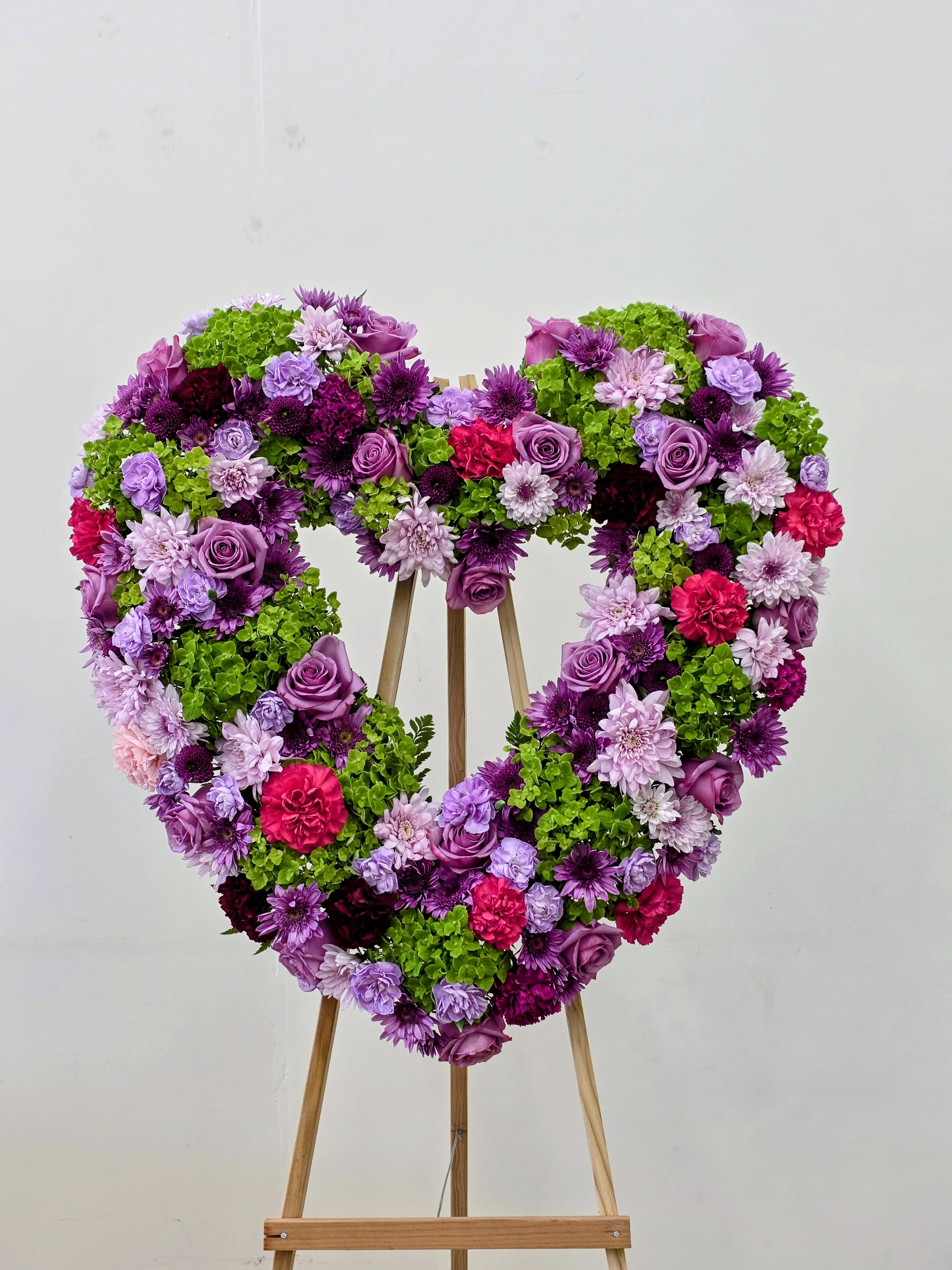 Lasting Love  - A large heart-shaped wreath bursting with purple roses, pink carnations, lavender and pink chrysanthemums and bright green hydrangea clusters-this standing floral tribute is made to make someone feel deeply remembered or celebrated. As a heart-shaped flower arrangement for a memorial, celebration of life, anniversary, or any love-themed tribute, it speaks clearly: the person you're honoring is cherished and unforgettable.  Perfect for placing beside a casket, at a service, or as a prominent statement at an anniversary or remembrance event, the easel-mounted heart makes the sentiment visible and unmistakable. Because it's designed for display, minimal care is needed-position it where it will be seen, and the dense construction will hold its shape and color throughout the event. 