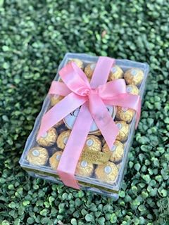 Large Chocolate Box -Pink - A glossy clear plastic gift box tied with a wide pink ribbon makes this Large Chocolate Box - Pink a cheerful, giftable flourish for birthdays, Mother's Day, or a sweet "just because" surprise. The neat rectangular presentation feels both refined and playful-perfect when you want the person you're thinking of to feel seen and celebrated, whether it's a romantic gesture or a thoughtful extra to pair with flowers.  Inside, the box is filled with gold-wrapped chocolates whose metallic shine contrasts beautifully with the soft pink ribbon and hints of red, black, yellow, and other tones visible through the clear top. The tidy, compact size makes it an ideal add-on to bouquets or a standalone token: small enough to tuck into a gift bag, large enough to make an impression. Packaged ready to gift, it arrives clean and presentable so the recipient experiences that little moment of delight the instant they lift the ribbon.