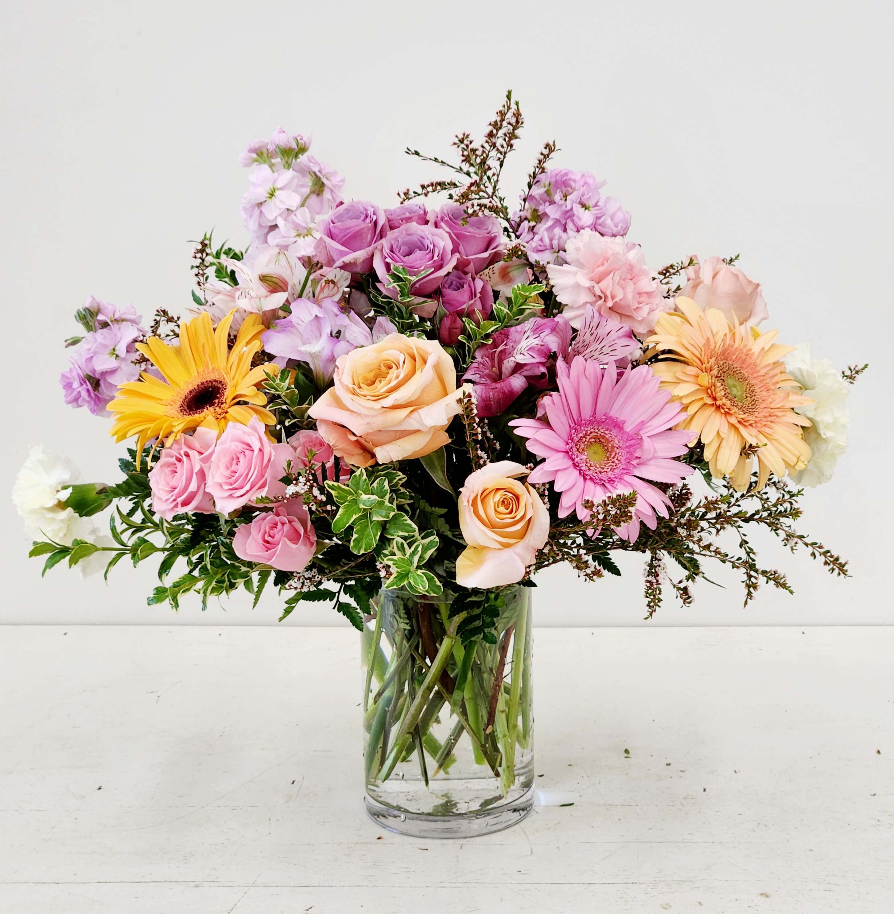 Whimsical - Soft swirls of pink, lavender, peach, and sunny yellow make this Whimsical flower arrangement feel like a garden picked at its brightest moment-perfect as a birthday bouquet, anniversary arrangement, or a cheerful spring gift. Roses and gerbera daisies form the eye-catching focal points so the person you're thinking of will first notice romantic blooms and joyful, open faces that say celebration and warmth.  This arrangement is designed to feel effortlessly festive for the recipient-she'll feel noticed, celebrated, and comforted by the gentle colors and lively shapes. Standard vase sizing offers a generous presentation; refresh water every two days, recut stems at an angle, and keep away from direct heat to extend the life of the blooms.