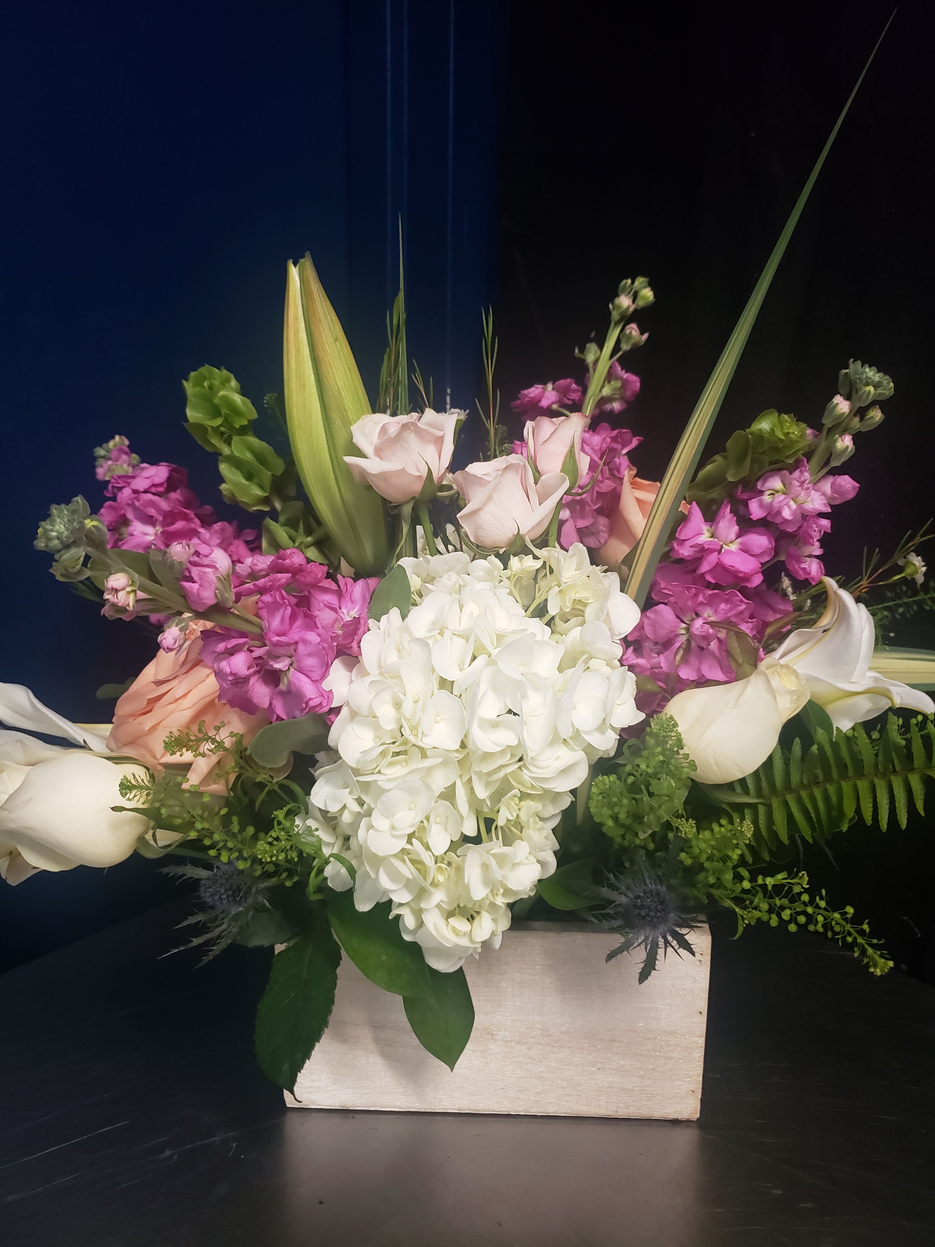 MOTHER'S DAY BOUQUES # 1-3 - A lush cluster of creamy white hydrangea anchors this Mother's Day flower arrangement, surrounded by blush and peach roses, white calla lilies, pink stock and soft spray carnations for a romantic, celebratory feel. Designed as a Mother's Day bouquet, it reads fresh and elegant-perfect for Mother's Day, a birthday, or to say thank you-so the person you're thinking of feels truly seen and celebrated the moment they receive it.  Each design uses seasonal pastels-white, pink, peach, purple and bright green foliage-for layered texture and movement. Options come arranged in either a low rectangular wooden vase or a tall clear glass vase: choose the rectangular wooden vase for a grounded, modern centerpiece, or the clear glass vase for a taller, garden-style look with blooms that extend outward. A lavender satin ribbon finishes the tulip-style version for a polished, gift-ready presentation. Care: keep flowers in fresh water, trim stems and refresh water every 2-3 days to extend vase life.