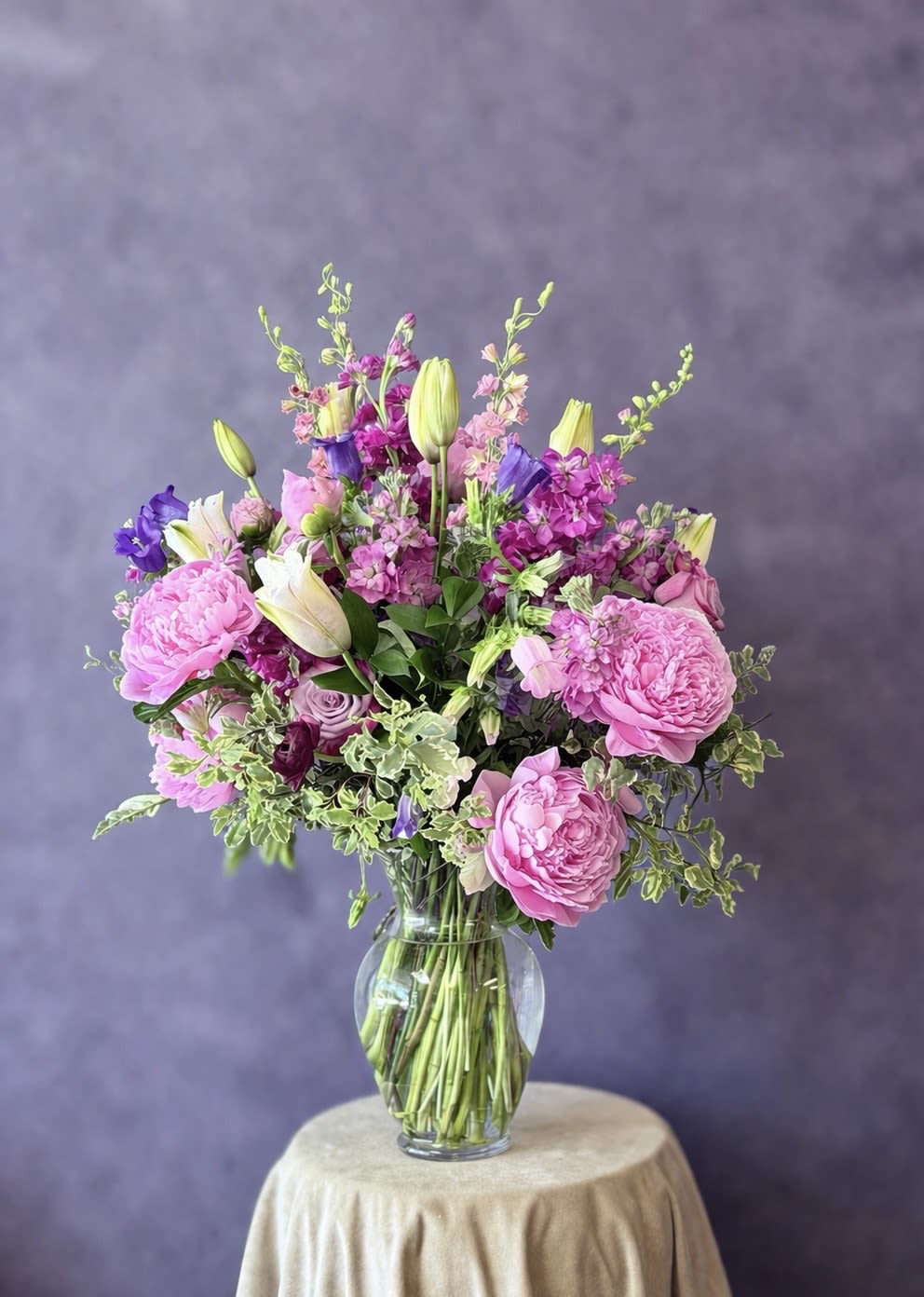 Purple Fantasy  - Soft lavender and vivid purple blooms mingle with bright pink gerbera daisies to create a gentle, uplifting flower arrangement perfect for a sympathy or funeral tribute or a heartfelt Mother's Day gift. This flower arrangement pairs airy stock and upright snapdragons with round chrysanthemums and lush roses so the person you're thinking of will feel comforted and seen; the overall palette of purple, lavender, pink, white, and green reads as both soothing and hopeful.  Two style options let you choose the mood: a loose, modern composition in a matte two-tone ceramic vase (beige top, muted green base) for a calm, contemporary display, or a fuller, rounded bouquet in a clear glass vase with peony-like blooms, roses, white lilies, and upright stock for a more romantic, garden-fresh look. Textures include soft stock and lisianthus, velvety roses and peonies, crisp snapdragons, and cheerful gerbera - arranged to provide visual height, fullness, and a fragrant presence that works well as a memorial piece, a thank-you, or a spring celebration centerpiece.