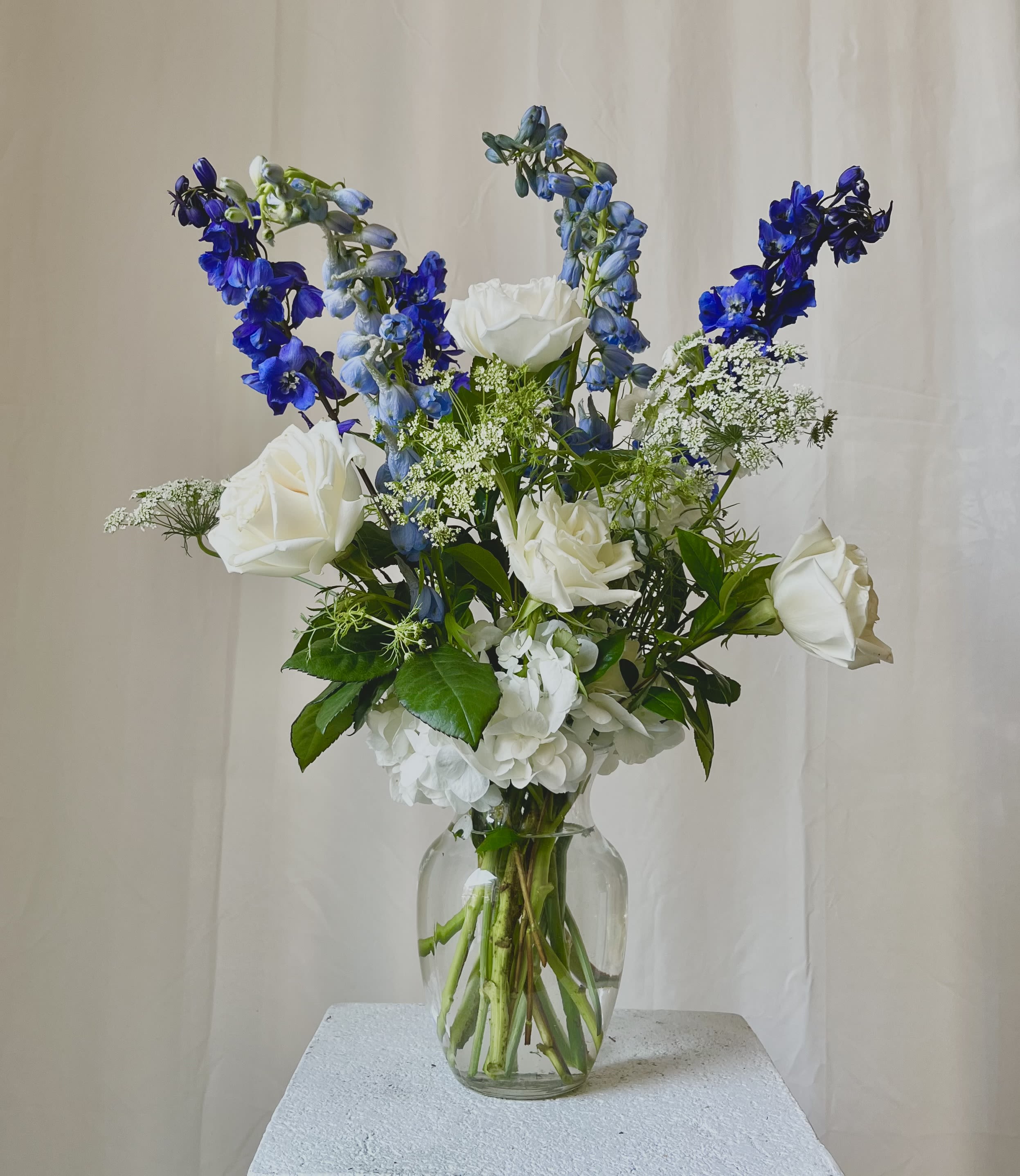 Tall arrangement of white roses and hydrangea with blue delphinium in a clear glass vase