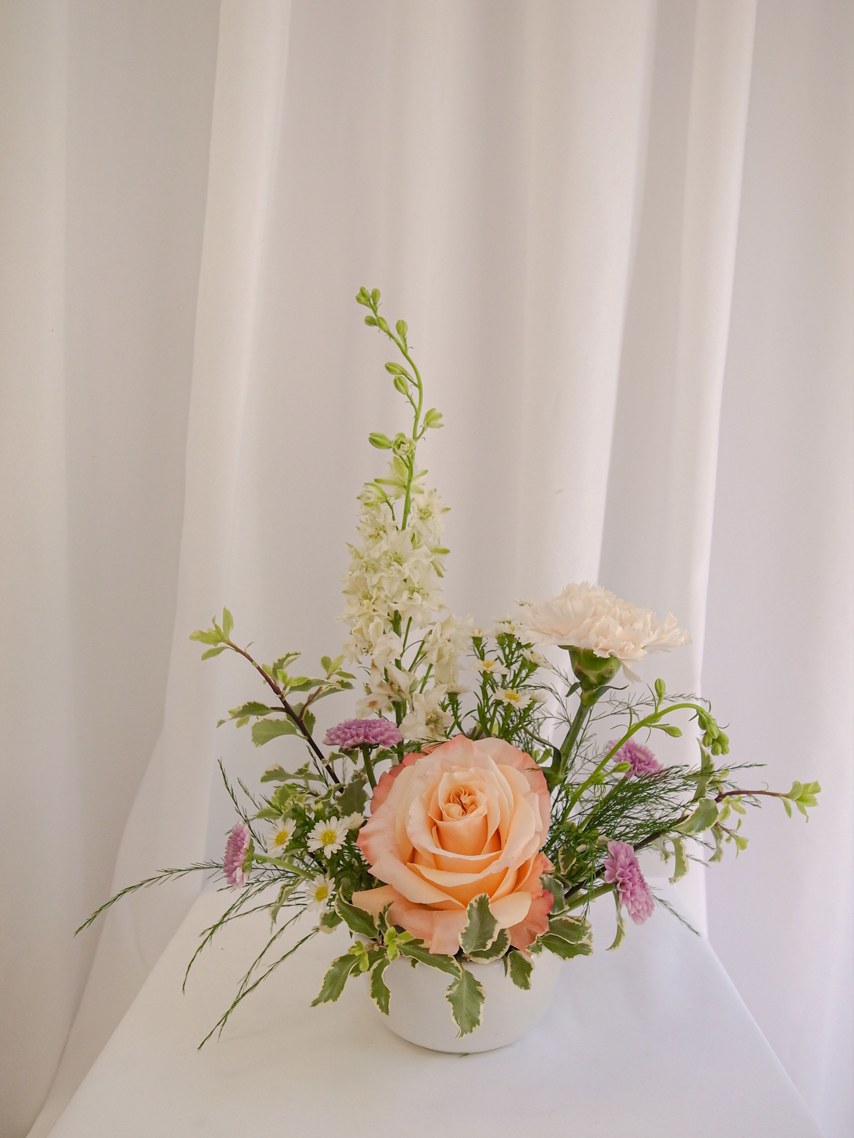Whispering Garden Mini - A delicate mini arrangement featuring a soft rose, airy white blooms, and fresh garden accents in a petite white vessel. Sweet and elegant—perfect for a thoughtful little gesture.