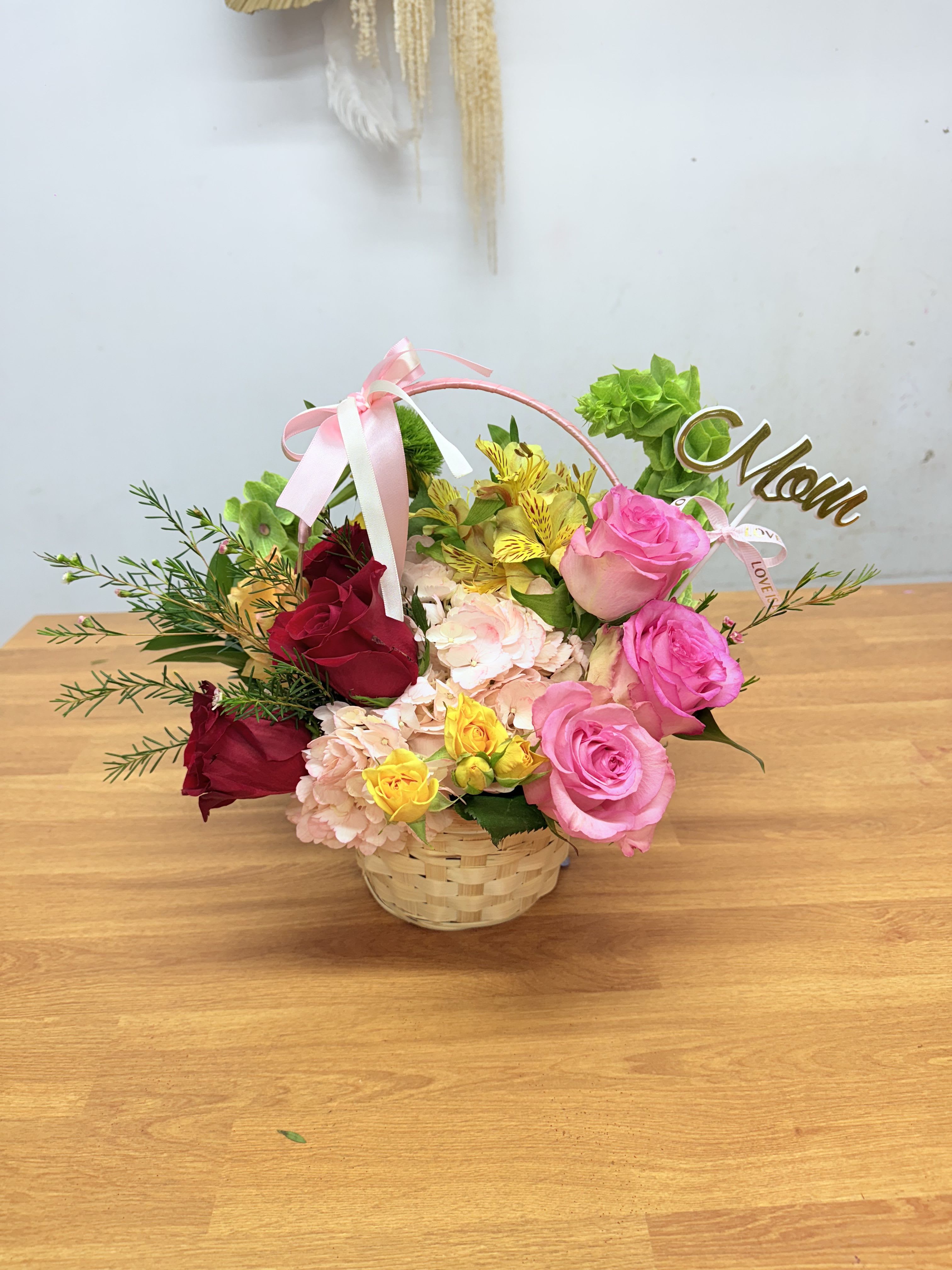 Mom pretty - Bright and cheerful, this Mom pretty flower arrangement is a colorful basket bouquet perfect for Mother's Day, birthdays, anniversaries, or anytime you want to say "I'm thinking of you." Pink and red roses take center stage to convey love and appreciation, while pops of yellow alstroemeria and soft white hydrangea clusters keep the mood joyful and fresh - the decorative "Love" pick and pink-and-white ribbon bow underline the affectionate sentiment.  Crafted in a small woven basket, the arrangement features rounded, abundant styling with airy greenery that gives a lively, hand-tied feel. Complementary carnations add soft texture among the roses and alstroemeria, creating a layered palette of pink, red, yellow, and white that looks full and inviting from every angle. The basket and ribbons make this piece gift-ready straight away.  Choose this for the mom, grandmother, or special woman who deserves to feel seen and celebrated - it reads as both intimate and festive, a simple way to brighten someone's day. Care is straightforward: keep in a cool spot away from direct sun and refresh the water as needed to prolong bloom life.