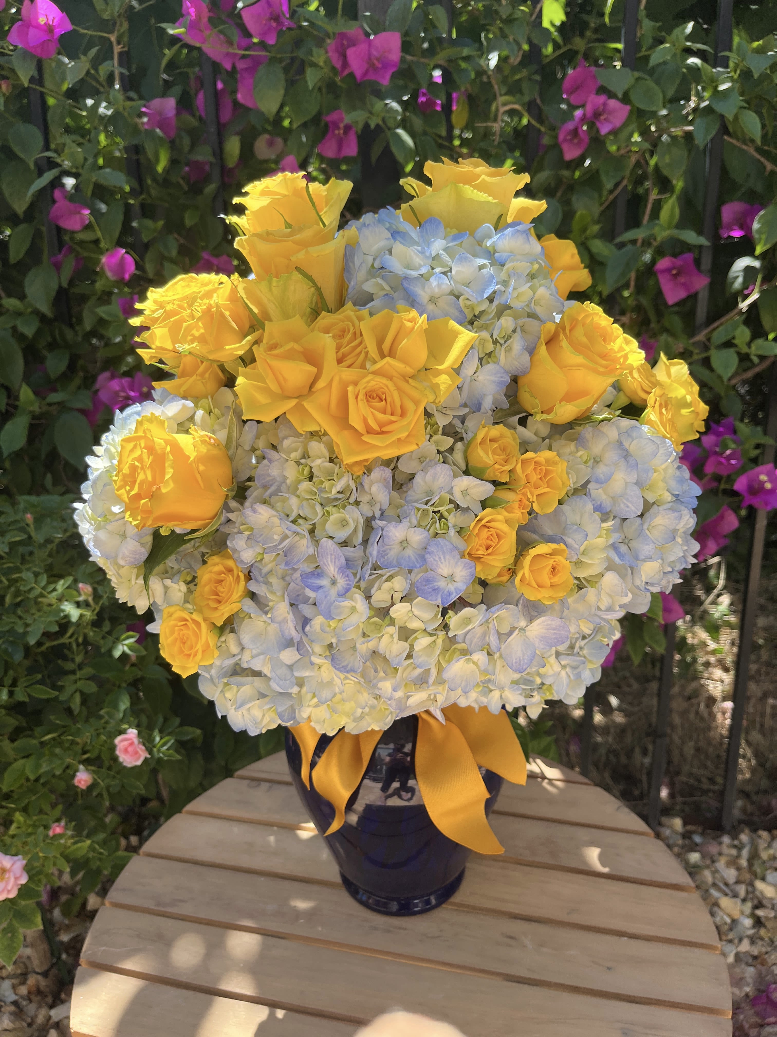 Yellow roses and blue hydrangeas in a dark vase with a yellow ribbon