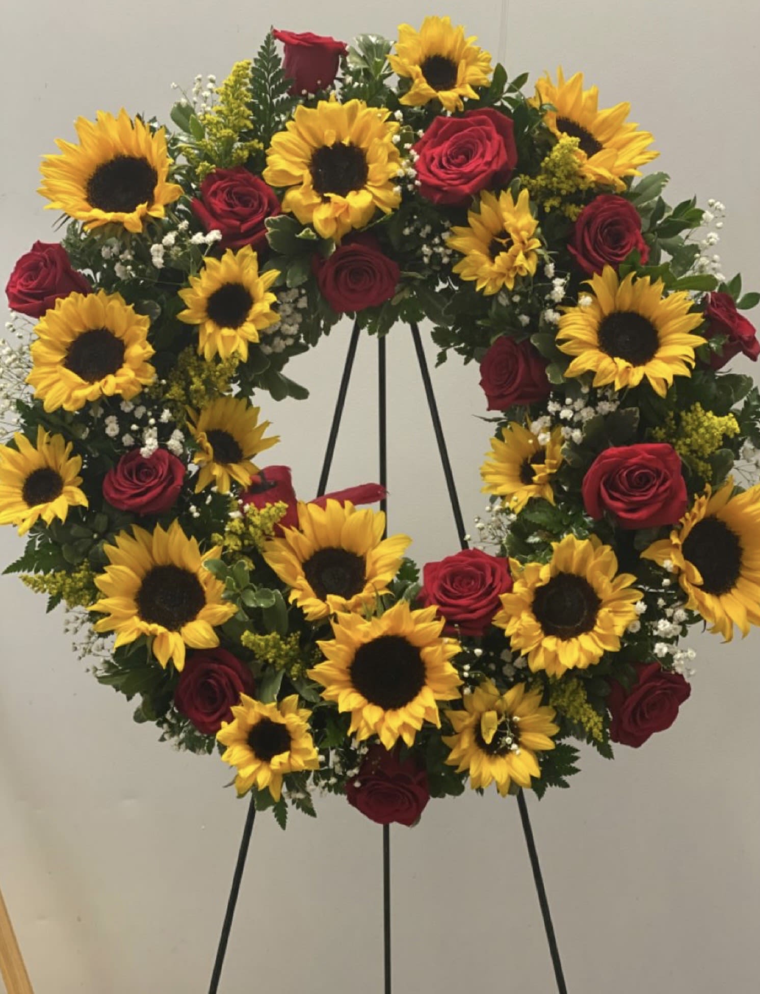 Golden Remembrance Wreath - Honor a life beautifully lived with our Golden Remembrance Wreath. This elegant sympathy arrangement blends radiant sunflowers with deep red roses, symbolizing warmth, respect, and everlasting love. Thoughtfully designed in a classic circular form, it represents eternity and the unbroken bond of memory.  Each bloom is carefully selected to convey comfort and heartfelt condolences during difficult times. The vibrant sunflowers bring a sense of light and peace, while the rich red roses express deep admiration and remembrance.  Perfect for funeral services, memorials, or graveside tributes, this wreath offers a dignified and meaningful way to say goodbye and celebrate a cherished life.  Let your tribute speak with grace, beauty, and sincerity.