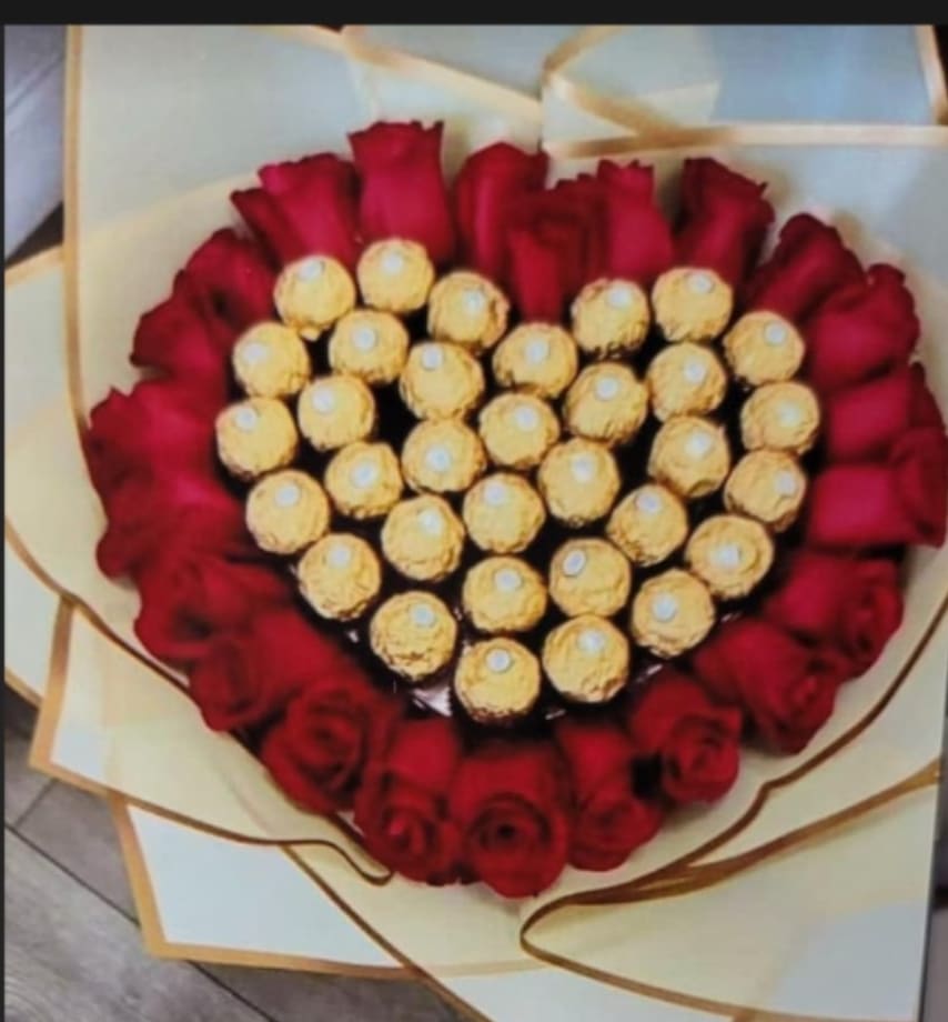 CHOCO HEART - A heart-shaped cluster of deep red roses ringed around a glittering center of gold-wrapped chocolates-this flower arrangement is a romantic bouquet designed for anniversaries, Valentine's Day, or any occasion when you want your person to feel adored. The red-and-gold contrast reads as both elegant and celebratory, so it works equally well as a surprise for a sweetheart or a special birthday gesture that says "I cherish you."  The design uses rich red roses arranged in a tight, symmetrical heart with Ferrero Rocher and assorted chocolate truffles densely packed into the center for an edible, eye-catching focal point. Cream-colored decorative wrapping paper with a thin ribbon and gold trim frames the heart like a hand-delivered gift, creating a polished presentation that travels well. The combination of velvety petals and shiny chocolate wrappers adds visual texture and makes an immediate impression.  Perfect for lovers, partners, or anyone you want to celebrate with a romantic touch-this arrangement sends a clear message of affection and indulgence. Order with standard sizing for a hand-held presentation; treat the roses gently, keep them in a cool spot out of direct sun, and enjoy the chocolates soon after delivery for best flavor.