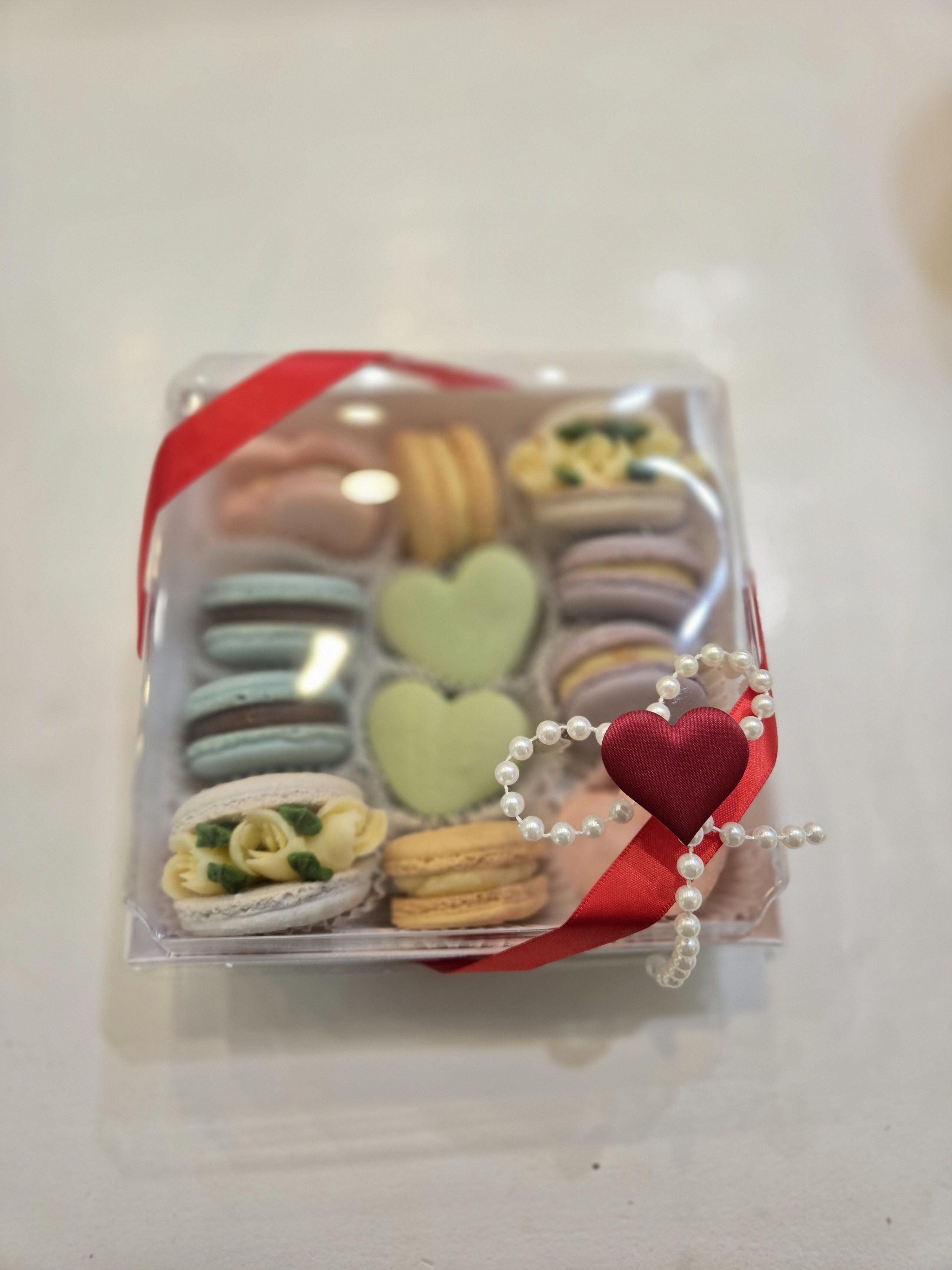 Artisan Macaron Gift Box - *** Please note: This item is available as an add-on only and cannot be purchased separately. It is designed to accompany one of our floral arrangements for a complete gift experience. ***  A stylish presentation of handcrafted sweets, designed to feel thoughtful and elevated. This clear gift box showcases an assortment of delicate French macarons in soft pastel tones, paired with heart-shaped cookies for a charming, romantic touch. Each confection is carefully arranged for visual appeal as well as flavor, creating a gift that feels both premium and personal. Finished with a red ribbon, pearl detail, and decorative heart accent, the presentation adds a subtle sense of luxury while keeping the focus on the quality of the treats inside.