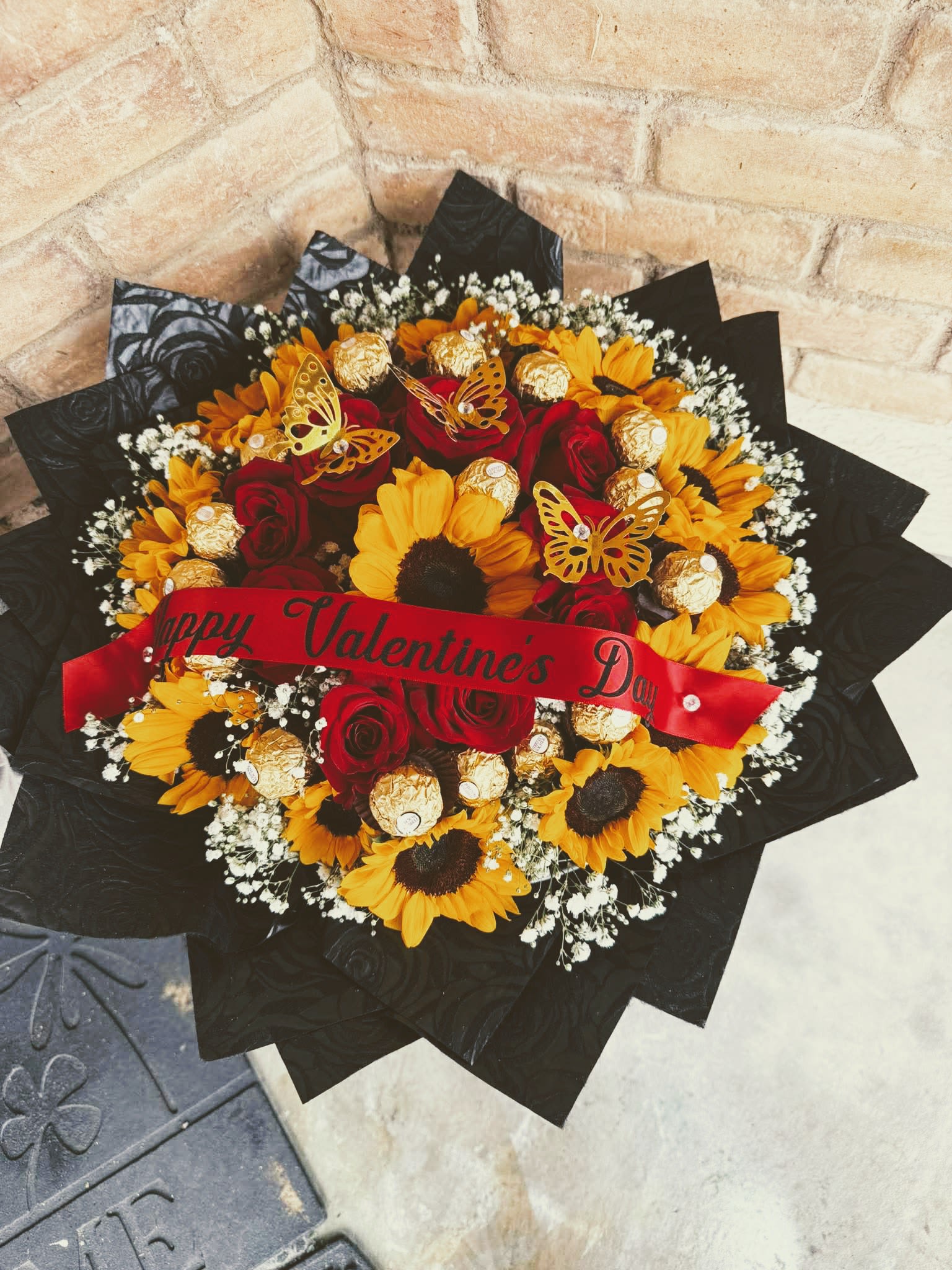 Ramo buchon sunflower and red rosas - A bold, rounded bouquet that pairs classic red roses with sunny sunflowers to create an unforgettable Mother day Valentine’s Day or anniversary flower arrangement. The contrast of deep red and vibrant yellow-brightened by delicate white filler blooms-makes the recipient feel adored and noticed the moment the bouquet is opened.  Crafted as a dense, "buchon" style hand-tied arrangement, the flowers are wrapped in dramatic black paper and finished with a red satin ribbon reading "Happy Valentine's Day." Gold foil-wrapped chocolate balls and decorative butterfly picks are nestled among the stems for an extra gift-like surprise, while the mix of textures-from velvety rose petals to coarse sunflower centers-keeps every angle interesting.  Perfect for a romantic surprise or to celebrate a special milestone, this arrangement sends a message of warmth and celebration. Size and care details: keep in a cool spot away from direct sunlight, refresh the water daily, and trim stems slightly if arranging into a vase to extend freshness.