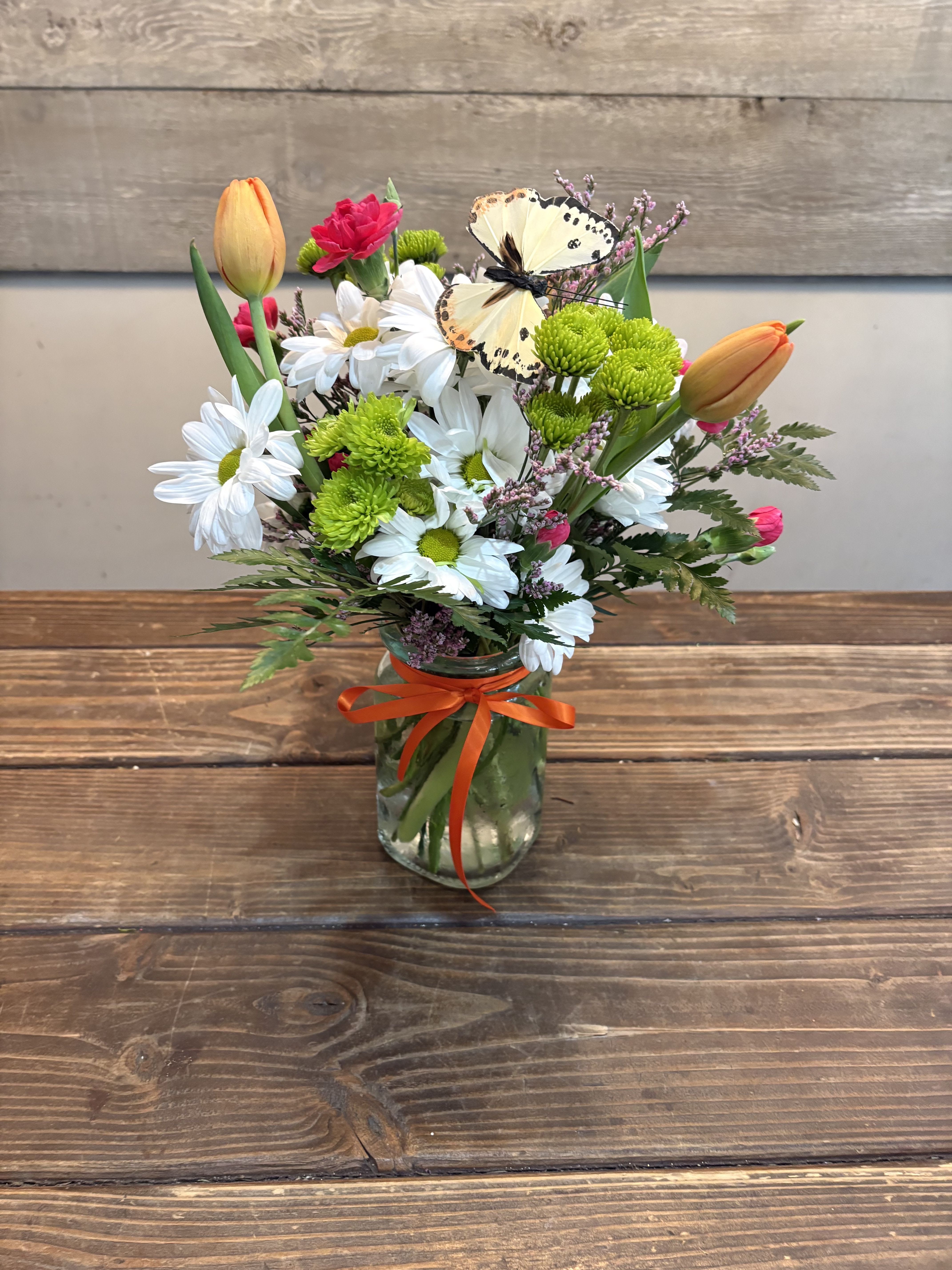 Sweet Daisy - A cheerful cluster of orange tulips and crisp white daisies topped with a playful decorative butterfly-the Sweet Daisy flower arrangement is a bright, springlike bouquet perfect as a birthday bouquet, a casual thank-you, or a seasonal celebration gift. The color mix of white, pink, orange, yellow and fresh green reads as joyful and lighthearted, so the person you're thinking of will feel cheered and noticed the moment they see it.  This compact arrangement is delivered in a clear glass jar (also shown in a compact vase option) tied with an orange ribbon for a finished, gift-ready presentation. It pairs white daisies with bright green button chrysanthemums, pink accent blooms and orange-yellow tulips for lively contrast and texture; a decorative butterfly perched near the top adds a whimsical focal point. The mix of soft petals and rounded mum buttons creates a fresh, garden-fresh look that fits casual and springtime occasions alike.  Sweet Daisy is sized for easy tabletop display-perfect on a kitchen counter, office desk, or bedside table-and arrives arranged and ready to enjoy. To keep blooms looking their best, refresh the water every couple days and trim stems slightly at an angle when you change it; this simple care will help the recipient enjoy the cheerful colors longer.