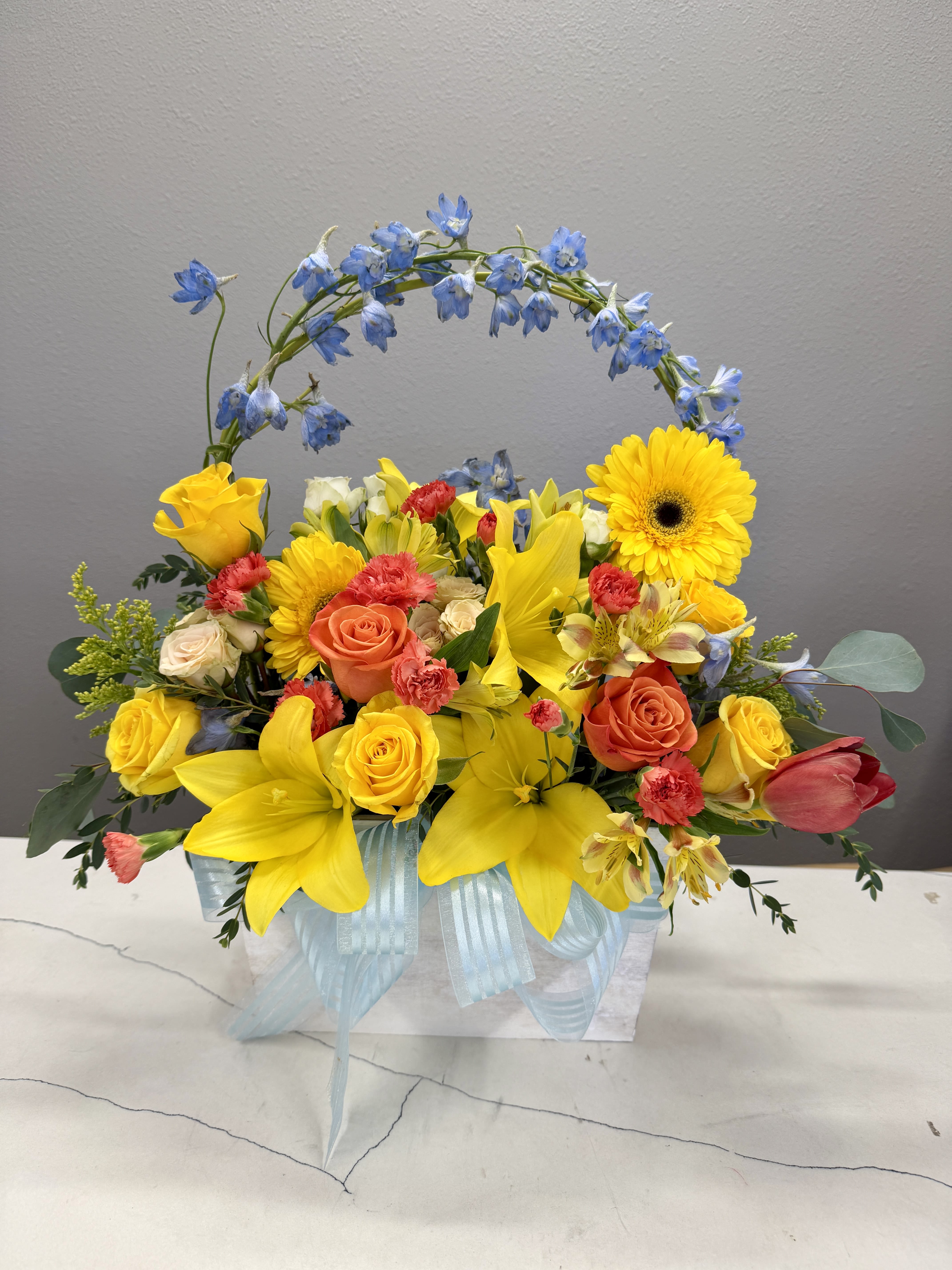 Sunlit Garden Basket - Bright, cheerful, and full of life, the Sunlit Garden Basket is a radiant floral arrangement designed to lift spirits and make any occasion feel special. This charming piece features a vibrant mix of golden yellow lilies and roses, warm orange blooms, soft peach accents, and delicate pops of coral and cream. A graceful arch of soft blue flowers adds a whimsical touch, creating a stunning garden-inspired silhouette.  Arranged in a keepsake container and finished with a soft blue ribbon, this design blends bold color with gentle elegance. Perfect for celebrations, thank-you gifts, birthdays, or simply bringing a bit of sunshine indoors, this arrangement delivers a fresh, joyful statement that feels both modern and timeless.