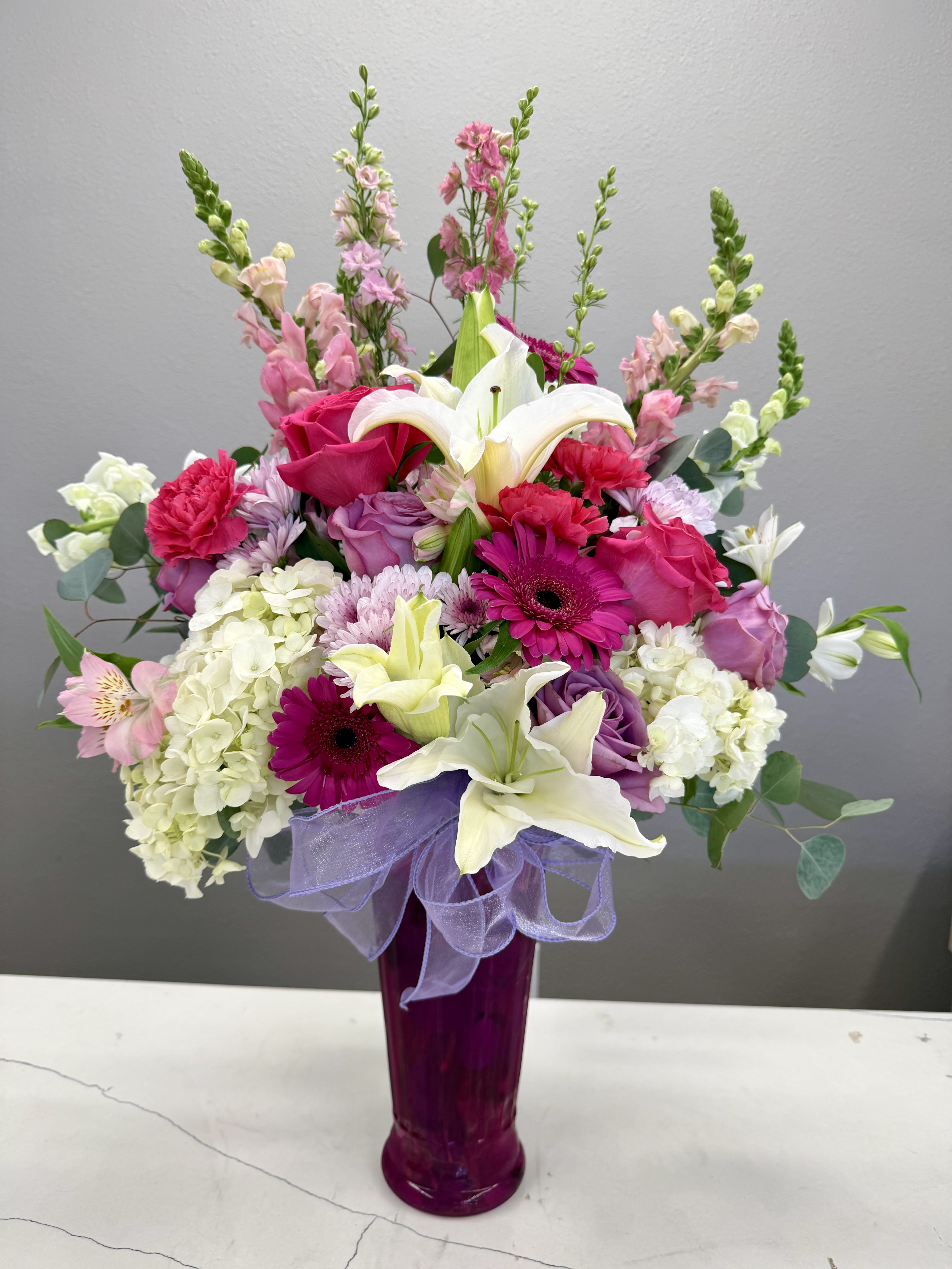 Petal Symphony Bouquet - Soft, romantic, and effortlessly graceful, the Petal Symphony Bouquet is a stunning arrangement that captures the beauty of delicate blooms in full harmony. Featuring lush white hydrangea, fragrant lilies, vibrant pink roses, and pops of magenta gerbera daisies, this design is accented with lavender tones and airy snapdragons for added height and texture.  Arranged in a sleek, deep pink glass vase and finished with a sheer lavender ribbon, this bouquet offers a perfect balance of sophistication and charm. Ideal for anniversaries, celebrations, or simply brightening someone’s day, it’s a timeless expression of love, appreciation, and joy.