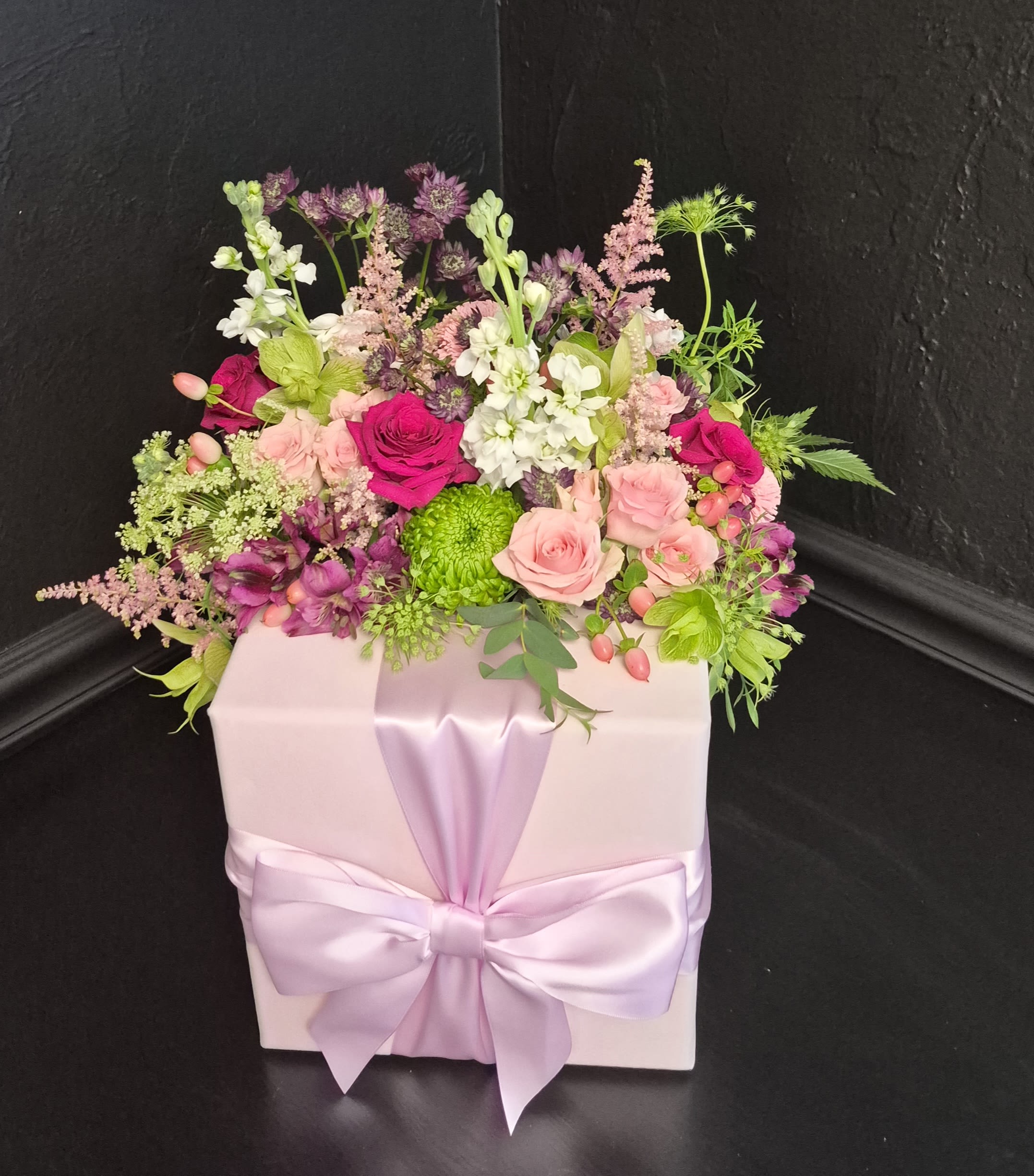Mother's Light - A dense cluster of blush and magenta roses with white stock and green daisies makes this gift-box arrangement feel like a secret garden in bloom - perfect as a birthday bouquet, anniversary flowers, or a Mother's Day surprise. The compact, romantic presentation rises with airy snapdragons and branching stems so the recipient sees layers of texture and color the moment the box is opened.  Carefully arranged in a pale pink gift box tied with a wide satin ribbon, the mix includes roses, stock, dianthus and other seasonal filler blooms in pink, purple, white and bright green accents that keep the palette fresh and lively. The box-mounted design creates a polished, celebratory look that travels well and needs minimal fuss - no vase required, simply place on a table and enjoy the fragrant, garden-style display.