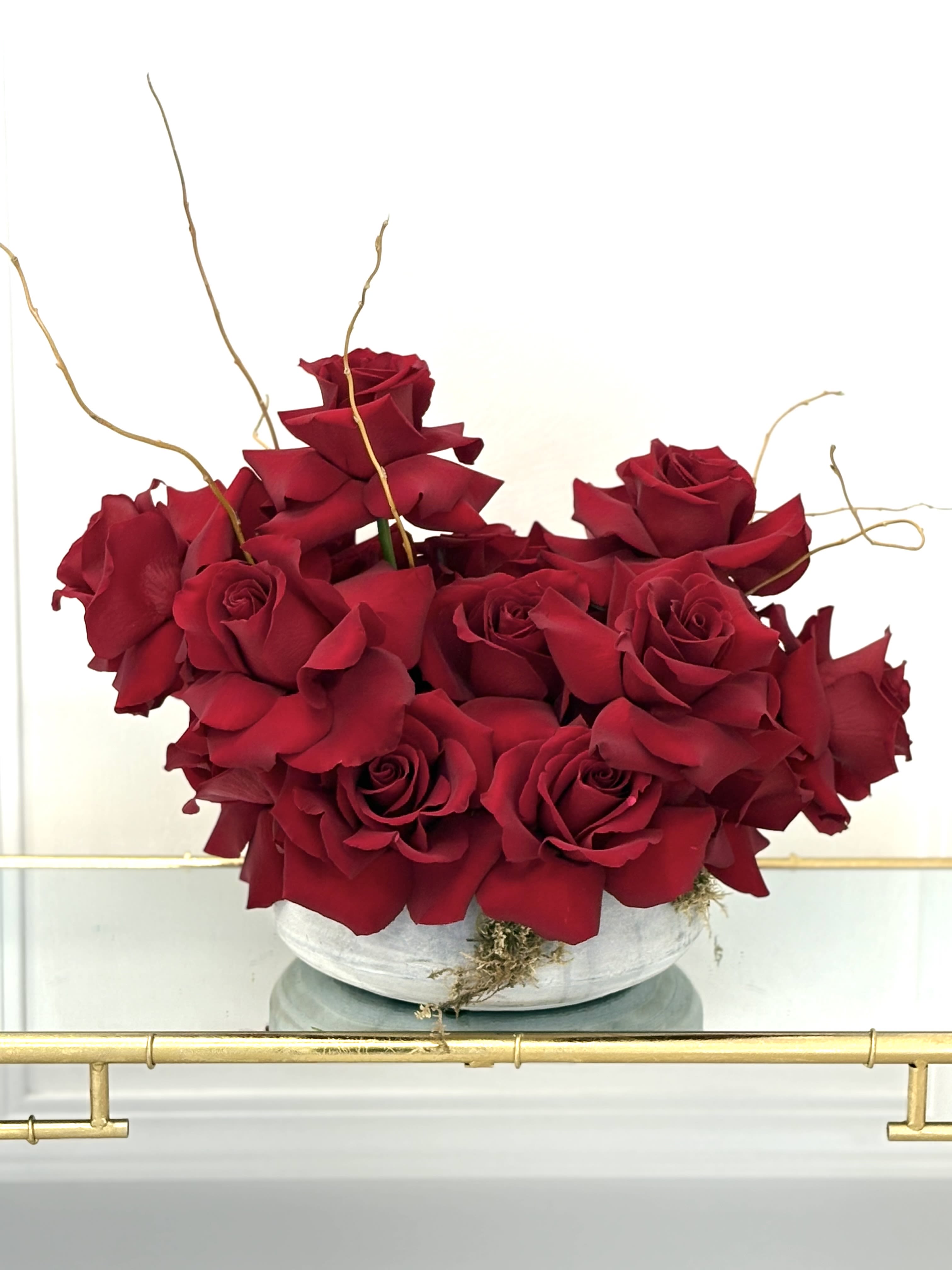 Low white bowl filled with clustered deep red roses and curly decorative branches