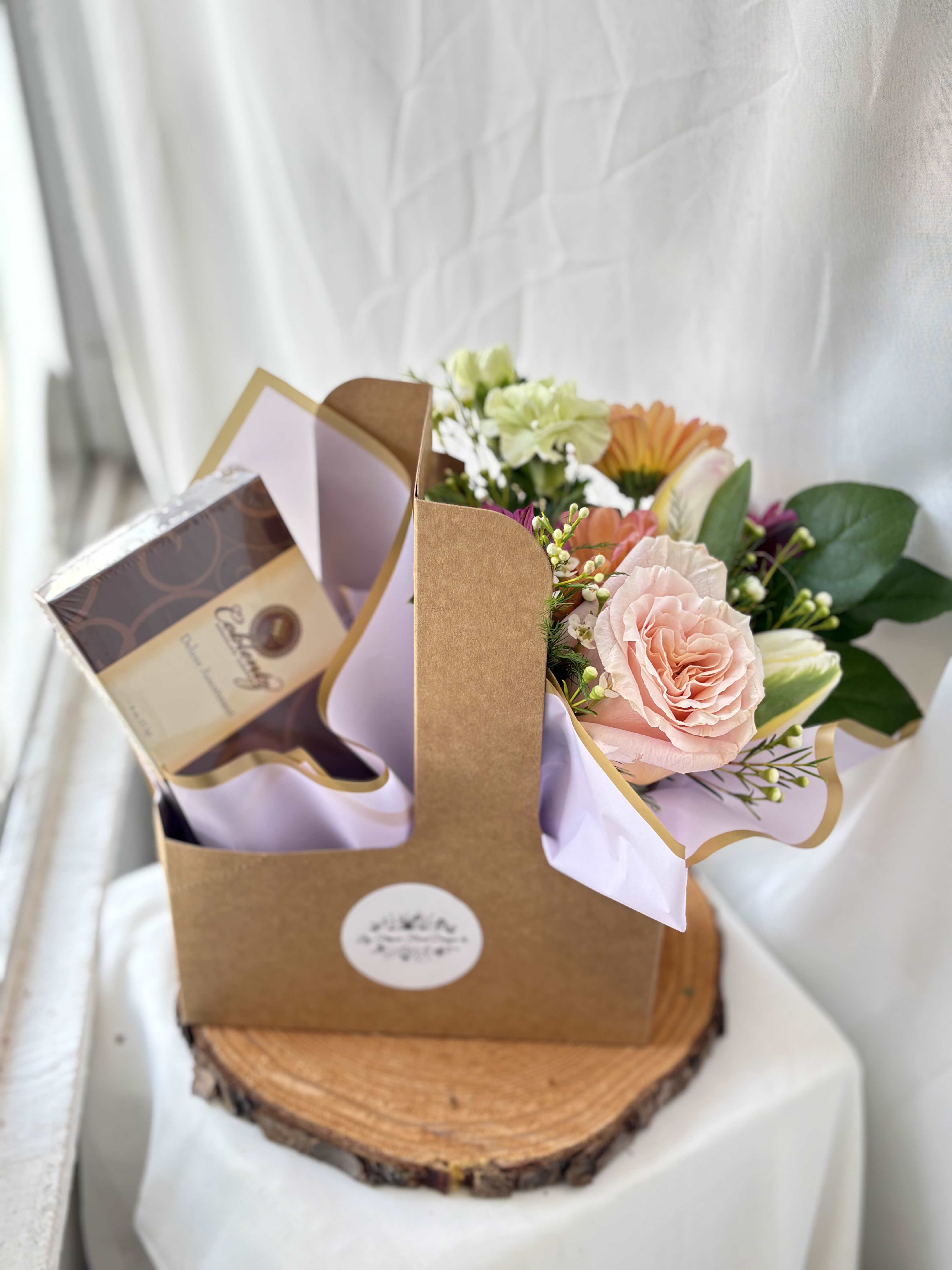 Gift caddy with pastel mixed flowers and a box of chocolates on a wood slice base.