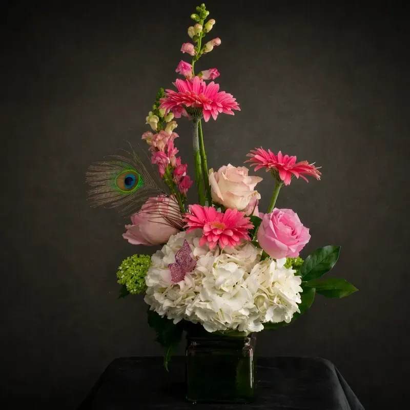 Gentle Grace - Soft and elegant design featuring a modern asymmetrical silhouette with delicate height and movement. This arrangement blends blush and soft pink roses with vibrant gerbera daisies, airy snapdragons, and a full base of white hydrangea for a lush, cloud-like foundation.  Accented with a touch of whimsy—a butterfly detail and a peacock feather—this piece offers a unique balance of playful and refined. Designed in a sleek glass cube, it’s perfect for adding a romantic yet contemporary statement to any space.  Each arrangement is handcrafted and may vary slightly based on seasonal availability while maintaining the overall style and color palette.