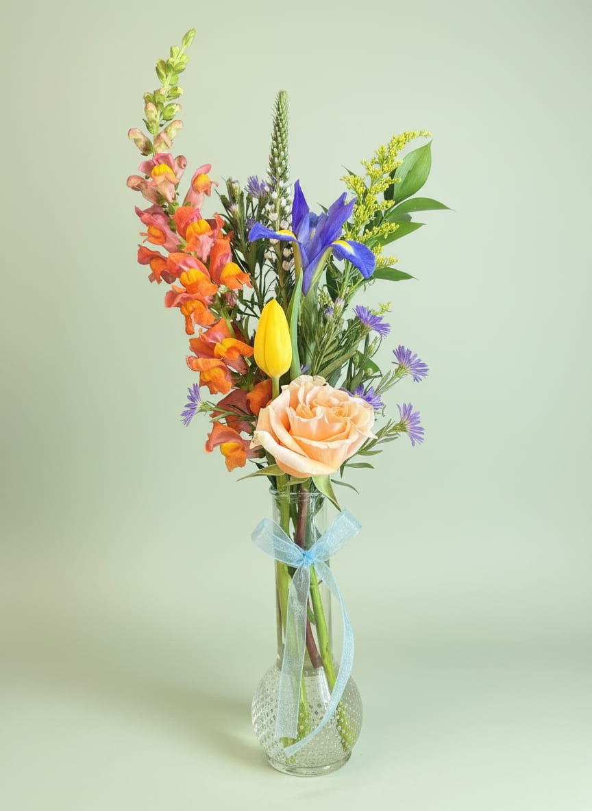 Bud Vase Designers Choice - A tall, airy bud vase arrangement that brings a fresh, garden-picked feel - perfect as a get well bouquet or a thinking-of-you gift. This mixed-color flower arrangement blends pink, purple, yellow and peach tones to create an uplifting focal point the recipient will notice right away; it also reads beautifully for Mother's Day or any spring celebration.  Carefully arranged in a clear glass vase tied with a pale blue ribbon, the designers' choice mix centers on a peach rose with bright yellow tulip accents, orange-pink snapdragons for vertical movement, and small purple asters and other seasonal blooms for texture. The loose, vertical composition feels lively rather than formal, giving each stem room to show its color and shape.  Made to brighten a bedside table, desk, or kitchen counter, this vase-sized arrangement is the kind of gift that helps someone feel genuinely seen and cared for. Designed with everyday gifting in mind, it arrives ready to display - just add fresh water and trim stems a bit if desired to extend vase life.