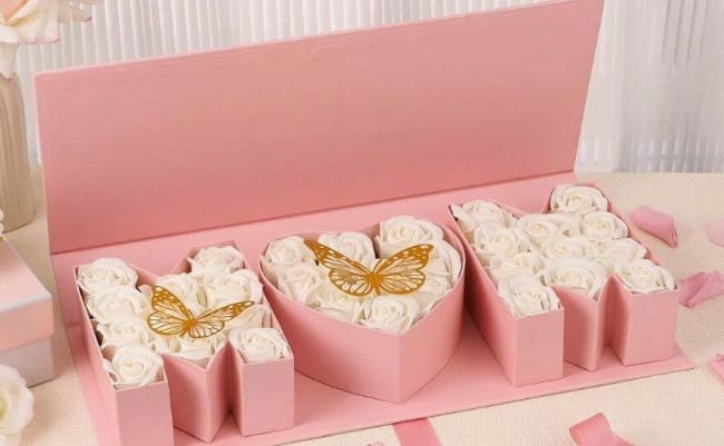 Mom Box White - A heart-shaped cluster of white roses set in a pink gift box creates an instantly romantic gift - perfect as a birthday bouquet, Mother's Day arrangement, or a surprise to say "I love you." The Mom Box White flower arrangement pairs white roses with soft pink and yellow accents and is designed to read as a celebratory, feminine present that makes the person you're thinking of feel truly seen and cherished.  The roses are arranged into three distinct sections inside a low pink presentation box: a central heart of tight white blooms flanked by two block-letter style clusters, giving the piece a sculpted, modern look. Tiny gold butterfly picks rest atop the rose clusters, adding a delicate metallic shimmer and a whimsical touch to the soft palette. Compact yet eye-catching, the box arrangement is easy to display on a table or desk and arrives ready to gift - no vase required.  This is the kind of gift that marks milestones and everyday moments alike: for moms, partners, or anyone who loves classic roses with a feminine twist. To keep the roses fresh, place the box in a cool spot away from direct sunlight and mist lightly if needed; sizing and water details are built into the presentation for hassle-free enjoyment.