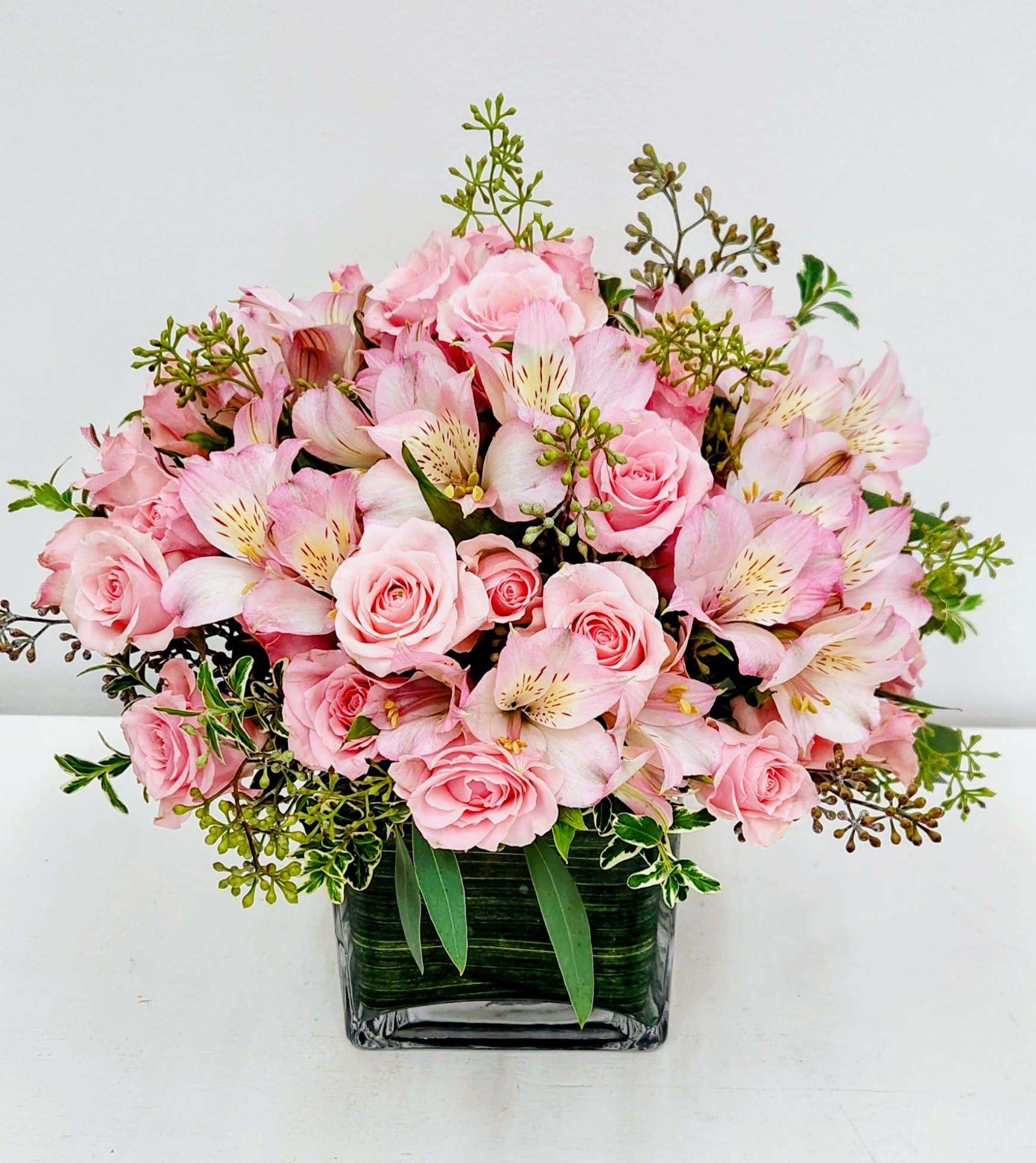 Long Beach Charms - A cluster of soft pink roses and pale pink alstroemeria creates a romantic, gently textured centerpiece perfect for a birthday bouquet, mother's day gift, anniversary flowers, or a thoughtful "I'm thinking of you" gift. The pastel pinks and fresh greens read as calm and celebratory, so the person you're sending it to will feel seen and warmly remembered the moment this arrangement arrives.  Dense, rounded blooms are arranged in a clear square glass vase with a few upright sprigs and variegated foliage to add height and movement. The medium-sized tabletop design fits well on a dining table, desk, or bedside table and highlights the creamy rose petals against the delicate alstroemeria. Simple care-fresh water in the vase and trimming stems every few days-keeps the arrangement looking its best for the recipient through the week.