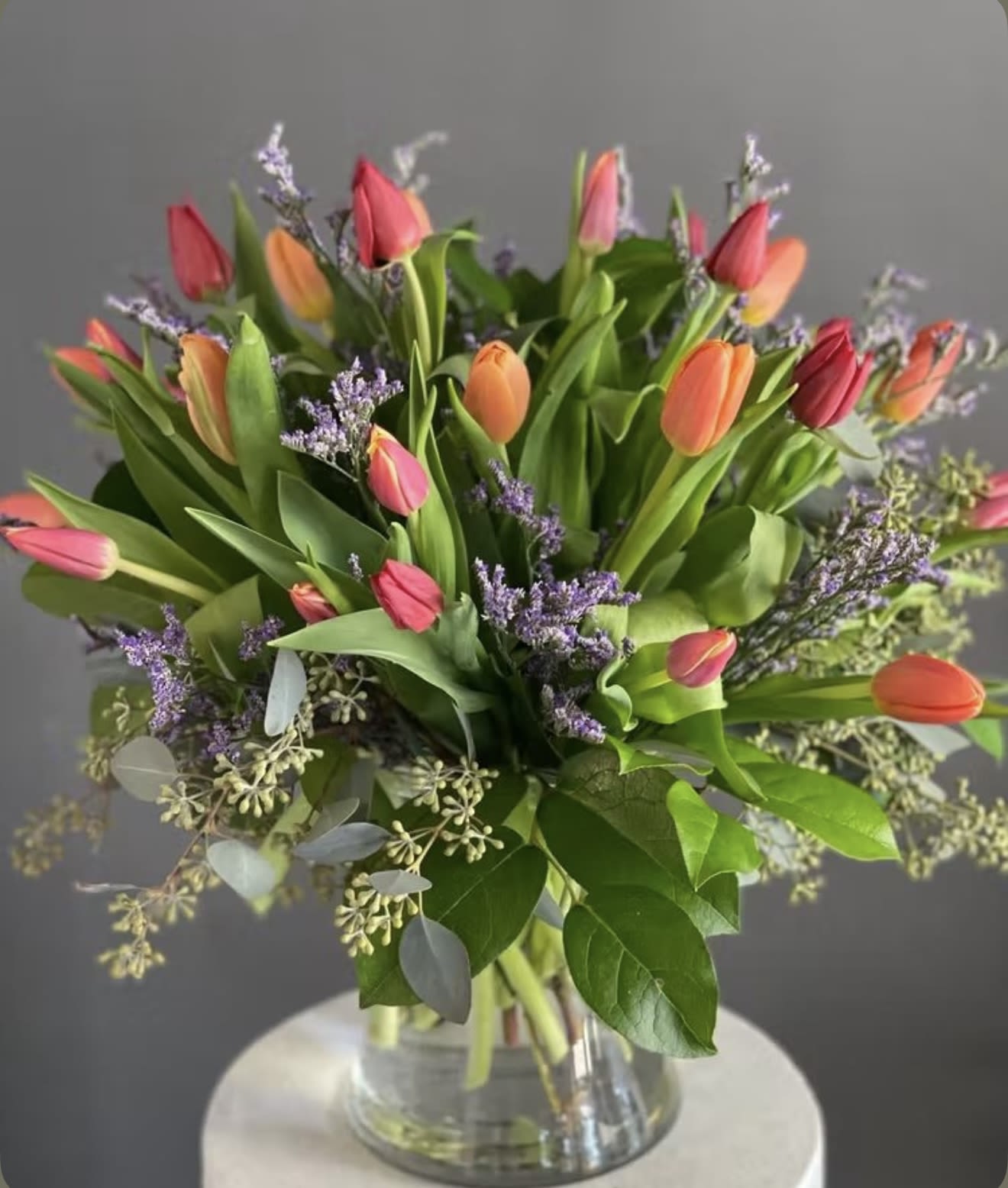 Tulips for a Mother’s  - A lively cluster of multicolored tulips creates an immediate fresh-spring impression-the perfect flower arrangement for a mother's birthday, Mother's Day, or a cheerful thank-you. Pink, coral, orange, purple and white tulip blooms spill from a clear glass vase with glossy green foliage and airy purple fillers, so the person you're sending it to will feel brightened and genuinely seen by its garden-fresh energy.  This arrangement pairs tightly gathered and looser, garden-style stems for a layered, natural look. Tulips in soft pinks, coral and orange sit alongside white and purple blooms, all complemented by fine filler flowers and broad green leaves; stems are visible through the clear cylinder for a clean, understated presentation. Sized for everyday gifting, it's an easy pick for birthdays, spring celebrations, or simply saying "thank you"-refresh water every few days, recut stems if desired, and enjoy the lively color mix that lasts while tulips are in season.