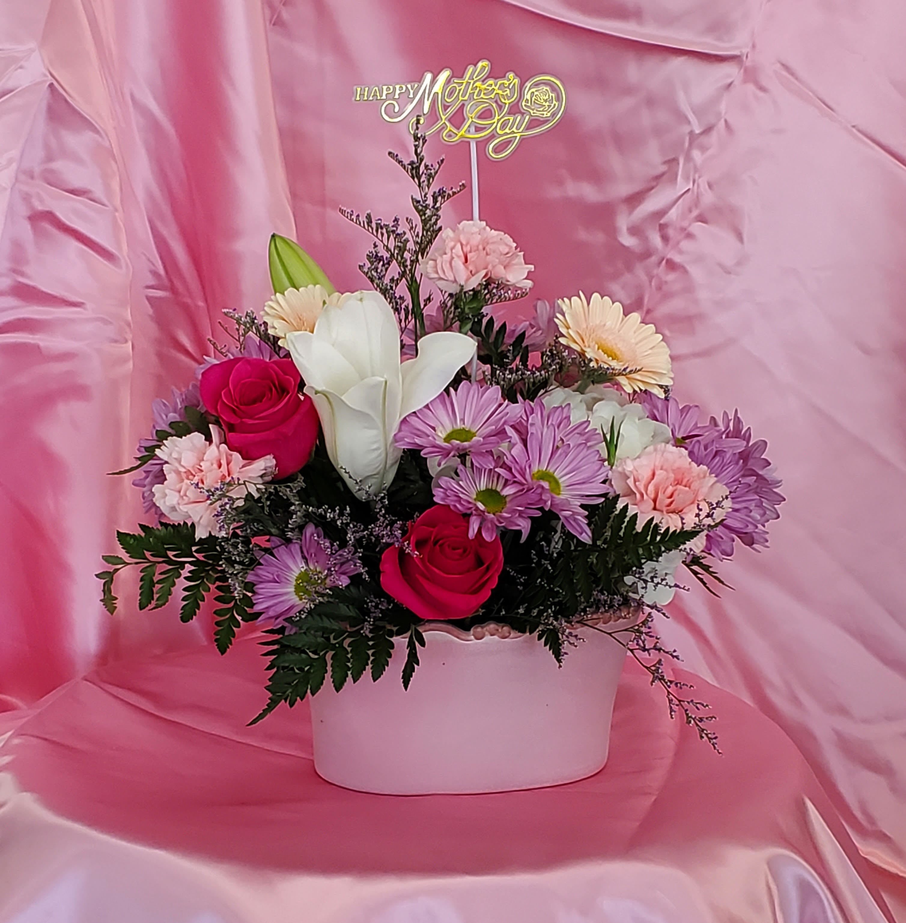 Mother's Day Garden Flowers  - Bright pink roses and soft white lilies take center stage in this Mother's Day garden-style arrangement, a cheerful floral gift designed to make the person you're thinking of feel truly cherished. The mix of roses, pale peach gerbera daisies, lavender aster (daisy mums) and petite pink carnations creates a layered, romantic look that reads as both joyful and elegant-perfect for Mother's Day, a birthday, or a thank-you moment when you want her to feel special.  Arranged in a low white ceramic container, the bouquet balances rounded blooms with daisy-like textures for a full, garden-fresh silhouette. The palette of pink, purple, white and green keeps the presentation light and feminine, while a gold "Mother" topper rises above the flowers to give the gift an instantly personal, celebratory touch. The compact design works well for tabletop or mantle display and arrives ready to enjoy.  This arrangement is especially suited for a mom, grandmother, or maternal figure who delights in soft, fragrant blossoms and cottage-garden charm. For best enjoyment, trim stems and refresh the water every few days to extend vase life; sizing is ideal for close-up display where the layered textures and colors can be appreciated.