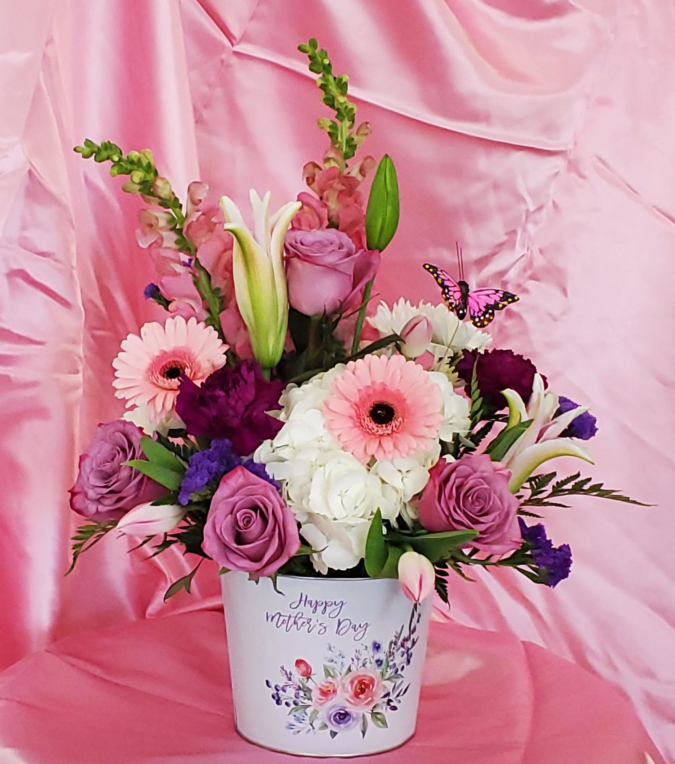 Bucket of Blooms  - Lavender roses and pale pink gerbera daisies mingle with billowy white hydrangea and elegant white lilies in a soft, spring-ready flower arrangement perfect for Mother's Day or a cheerful birthday surprise. Tall pink snapdragons add vertical drama while touches of purple and a few deeper-toned blooms give contrast against the pastel palette, so the person you're thinking of will feel celebrated and seen the moment this bouquet arrives.  Arranged in a white metal container printed with a Mother's Day message and floral artwork, the ensemble arrives gift-ready with a decorative butterfly pick for a playful finishing touch. The mix of textures - velvety roses, crisp gerbera centers, lush hydrangea, and stately lilies - creates a polished yet romantic display that brightens a kitchen table or entryway. Order now for spring delivery and enjoy simple care: keep water fresh and trim stems for longest enjoyment.