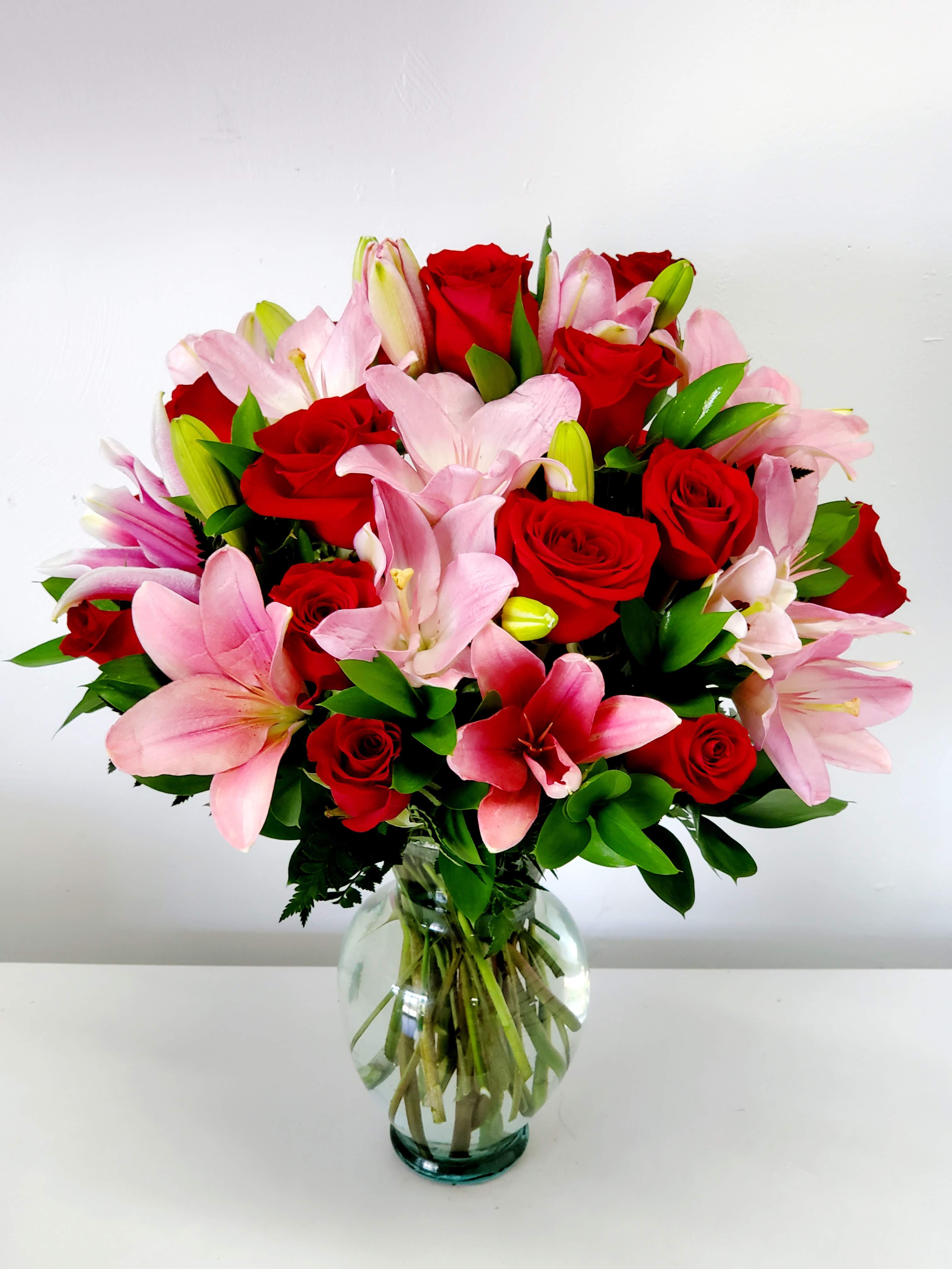 Enchanting  - Vivid red roses paired with soft pink lilies create a romantic, celebratory bouquet made to make the person you're thinking of feel deeply adored. Perfect as an anniversary bouquet or a love-themed gift, this flower arrangement also works beautifully for birthdays or "just because" moments when you want someone to feel seen and cherished.  The full, rounded design sits in a clear glass vase that shows the stems through the water for a simple, classic presentation. Saturated red roses provide rich color and texture while pale pink lilies open wide with soft, fragrant blooms; accents of green and touches of white brighten the composition and add contrast. Sized to stand out on a dining table or bedside table, this arrangement arrives ready to display and delight.