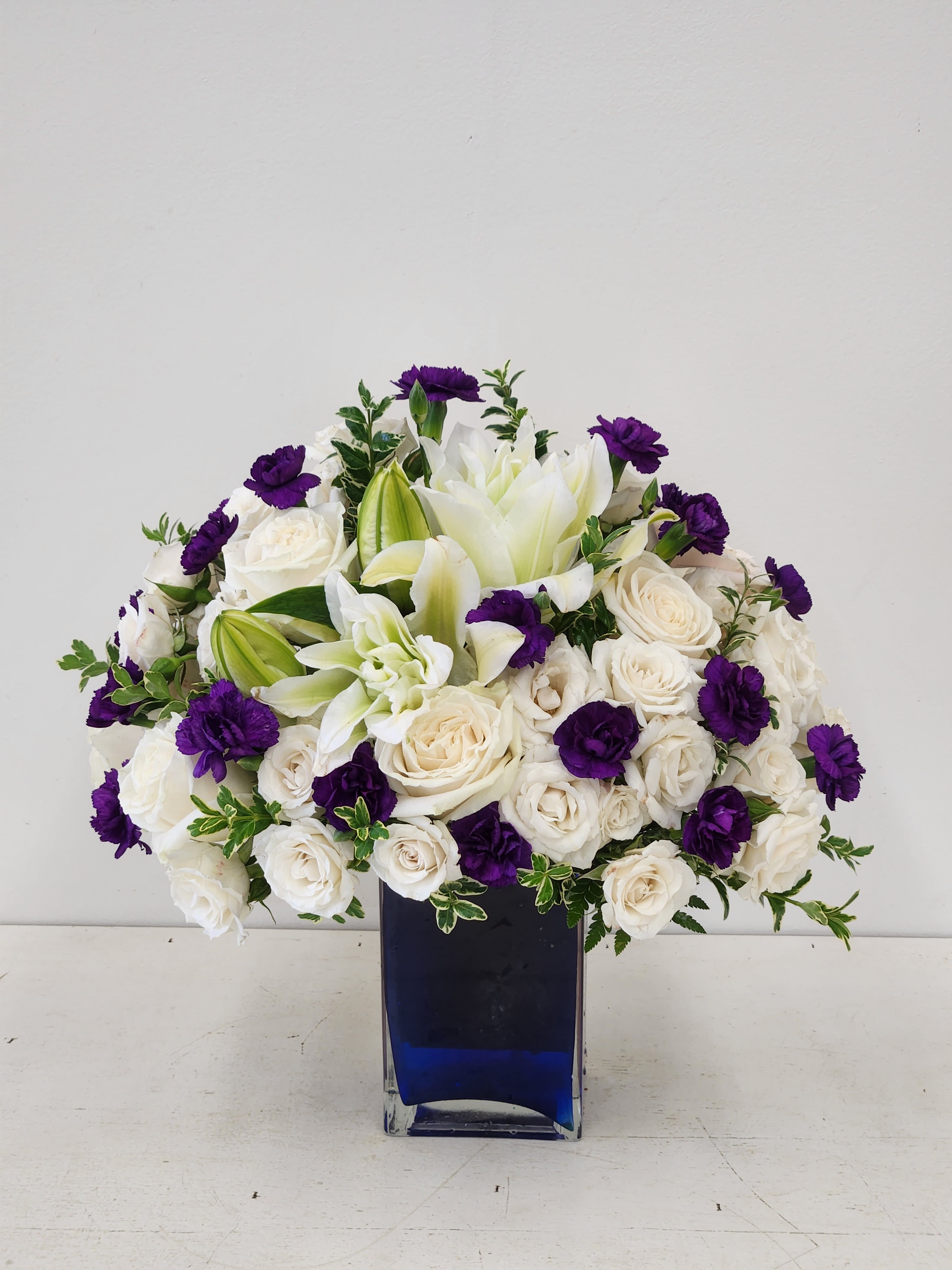 Violets in Blue - A refined mix of white roses and lilies punctuated by vivid purple carnations, this flower arrangement-perfect as a sympathy arrangement or for a formal wedding-creates an immediate sense of calm and grace. The rounded, full composition reads both ceremonial and comforting, the ivory roses and tall white lilies offering softness while the saturated violet carnations deliver a clear, uplifting accent that draws the eye.  Carefully gathered in a cool blue glass vase, the palette of white, purple, green, and blue feels balanced and serene. The lilies bring vertical movement, the roses add lush, layered texture, and the carnations contribute bright pops of color; together they form a dense, elegant bouquet that sits beautifully on an altar, reception table, or beside a bedside. Sized as a full, rounded arrangement, it arrives ready to display-simply place in a cool spot out of direct sunlight and change the water every two days to keep blooms fresh.