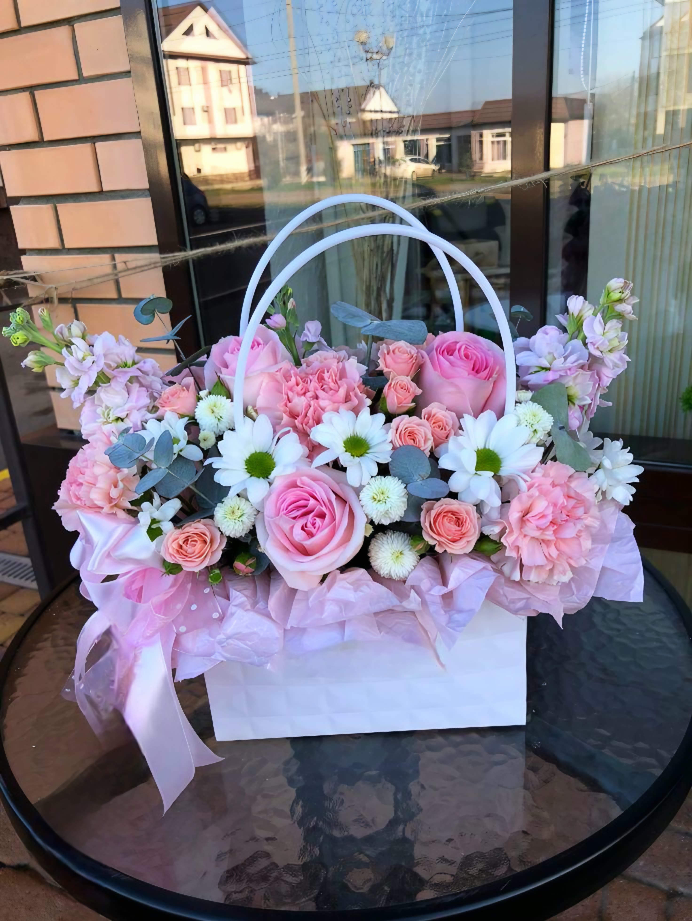 Mommy's Purse - A soft, pastel harvest of nostalgia-this flower arrangement in a white handled basket feels like a gentle hug for Mother's Day, a birthday, or any moment you want to say "I'm thinking of you." The rounded composition of large pink roses and clusters of spray roses reads as classic and romantic, while pale purple accents and blue‑green eucalyptus brighten the mix so the recipient feels both cherished and uplifted.  Carefully arranged with pink carnations and white daisy‑like blooms, the palette of pink, white, purple, and green creates a light, airy texture that suits spring celebrations and everyday gifting alike. The arrangement sits in a white basket lined with white tissue paper, finished with a pink ribbon around the handle for a polished, gift‑ready presentation. Sized to sit beautifully on a table or bedside, it's the kind of floral gift that makes someone feel genuinely seen and celebrated; simple care-fresh water and a cool spot-will keep the blooms looking their best.