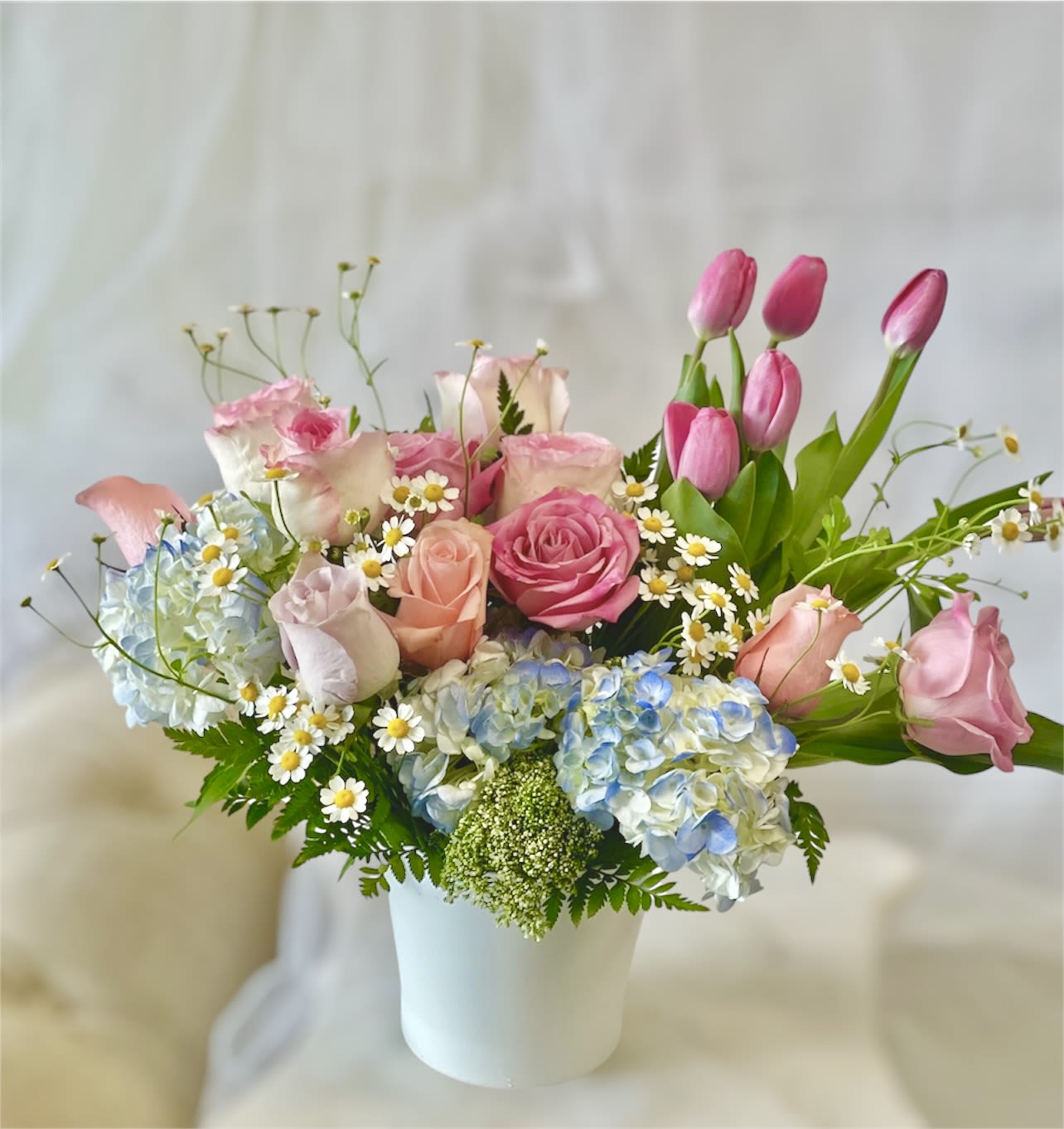 Pink roses and tulips arranged with blue hydrangeas in a white vase.