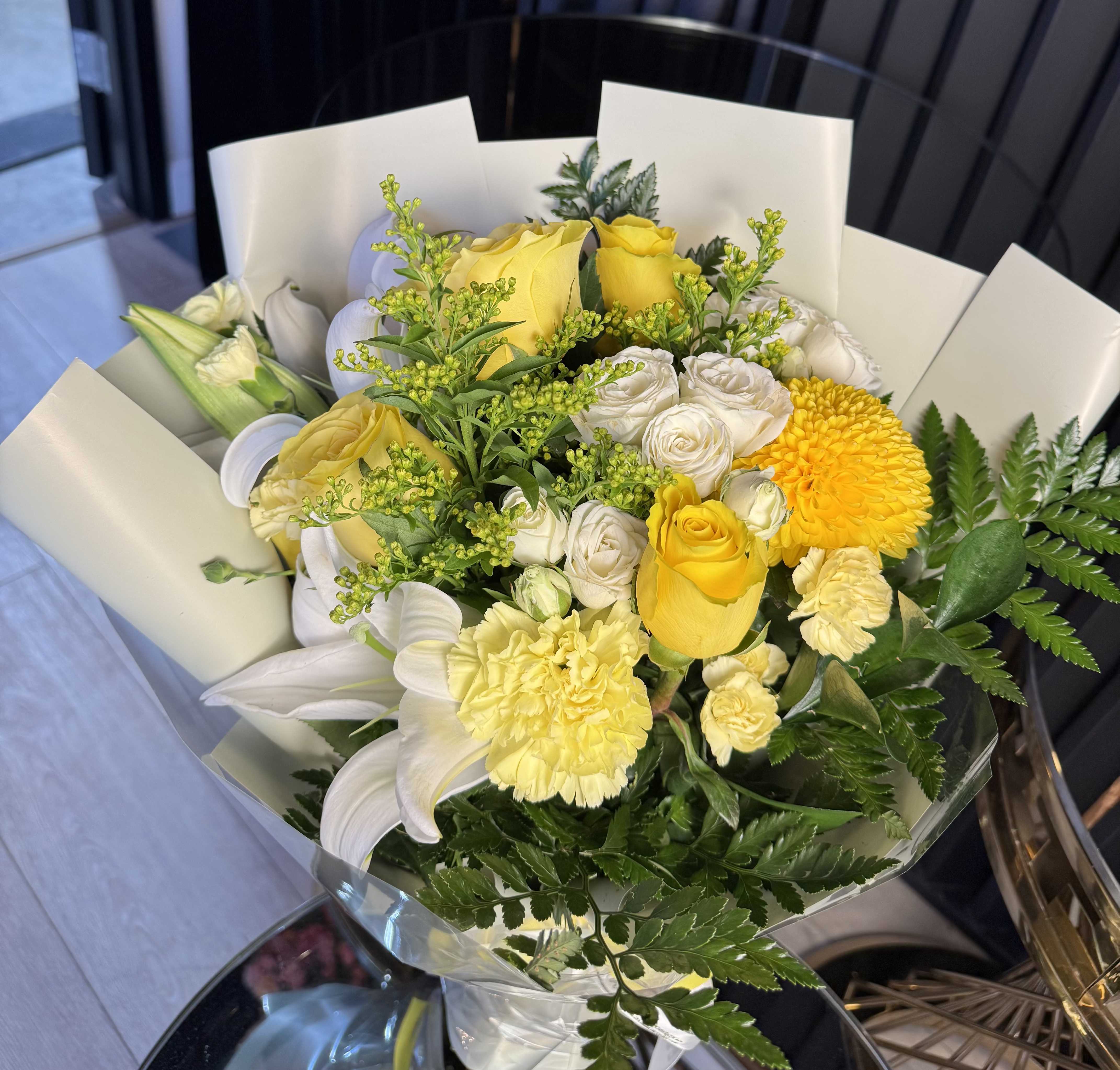 Sunny Serenity Bouquet - Radiate warmth and joy with this elegantly wrapped arrangement. Designed to mimic a sun-drenched garden, the Sunny Serenity Bouquet blends the classic romance of yellow roses with the striking architecture of white lilies. A focal yellow chrysanthemum adds a bold, modern texture, while soft carnations and sprays of goldenrod provide a lush, full finish.  Wrapped in a minimalist, layered ivory paper with a clean aesthetic, this bouquet is the perfect gesture for birthdays, "get well" wishes, or simply to say "you brighten my day.