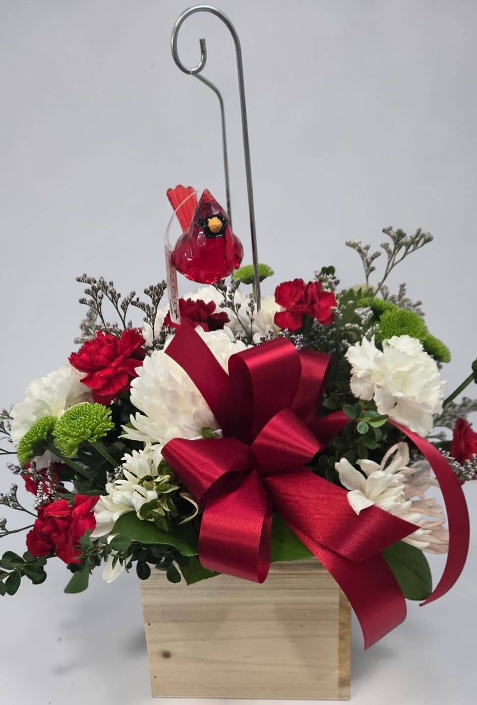 Cardinals Blessing by Art and Flowers -  Cardinal’s Blessing  This arrangement has a warm, nostalgic charm — the kind that feels like stepping into a cozy moment. A light wooden box cradles a lush mix of red carnations, white chrysanthemums, and green button mums, creating a classic red‑white‑green palette that instantly reads festive. Soft sprigs of greenery add texture and fullness, while a red satin bow anchors the design with a bold, celebratory touch.  Rising from the center is the standout detail: a red glass cardinal ornament, perched on a slender metal hook. It gives the whole piece a symbolic lift — cardinals are often associated with hope, remembrance, and warmth — making the arrangement feel both decorative and meaningful.  The overall effect is elegant, cheerful, and perfect for  simply brightening a room with something heartfelt.