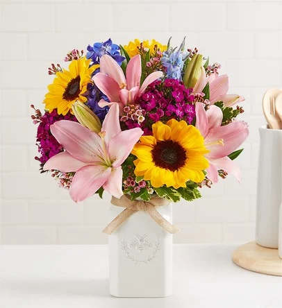Honey Bee Buzz - Get them buzzing with our charming bouquet! Sunflowers, Pink Asiatic Lily, Blue Delphinium, Fuchsia Dianthus, Pink Waxflower and Assorted Greens are gathered together for a mix of color and texture. Designed in our Sweet As Can Bee artisanal vase, featuring an adorable honey bee and raffia bow, it’s a gift that adds quaint beauty to any decor.  Arrangement with sunflowers, pink asiatic lily, blue delphinium, fuchsia dianthus, pink waxflower and assorted greenery Artistically designed in our Sweet As Can Bee vase, crafted in white ceramic, accented with an embossed honey bee crest and finished with a raffia bow; measures 4.13"L x 4.13"W x 7"H Premium arrangement measures approximately 19"H x 17"H Deluxe arrangement measures approximately 18"H x 16"H Standard arrangement measures approximately 17"H x 15"H Our florists hand-design each arrangement, so colors and varieties may vary due to local availability To ensure lasting beauty, Asiatic Lilies may arrive in bud form and will fully bloom over the next few days