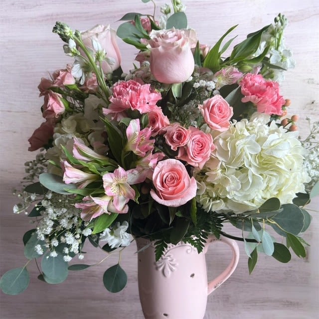 Perfect Pink Pitcher - Soft blush and pink roses mingle with pale hydrangea and pink carnations in a garden-style pitcher arrangement that reads romantic and relaxed-perfect as a Mother's Day, anniversary, or birthday bouquet. The flower arrangement and occasion are purposeful: send this as a Thinking of You or Thank You gift, or "just because" surprise that makes the person you're thinking of feel genuinely seen and cherished.  Alstroemeria, spray roses, stock, and airy white filler are tucked among eucalyptus-like green foliage for a layered, textural look that feels both lush and effortless. The flowers are arranged loose and abundant in a pale pink ceramic pitcher vase, giving the display a vintage, feminine charm that can be used long after the blooms fade. Available in a single carefully styled size, it arrives ready to display-no extra vase needed-so she can enjoy the scent, color, and thoughtful presentation right away.