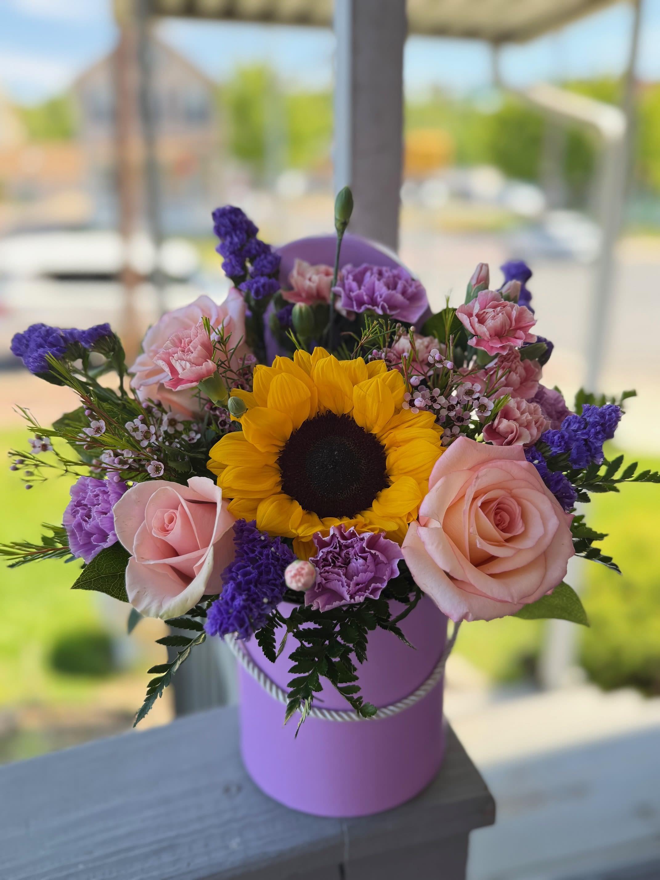 Violeta  - Round lavender hatbox with one bright sunflower, pink roses, carnations and purple fillers - compact giftable bloom mix