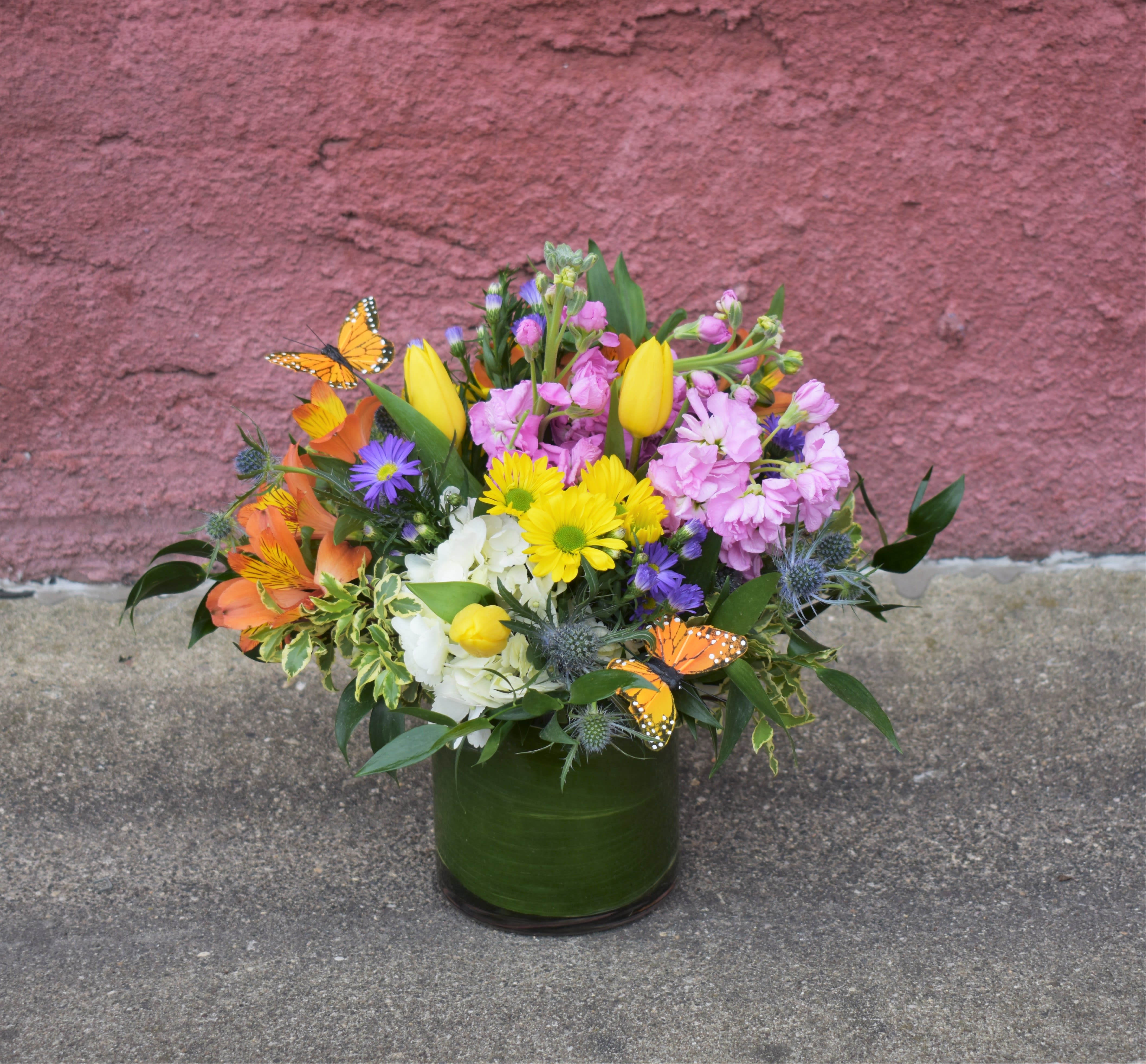 Wildflower - This bouquet is brimming with an array of colorful blooms! Orange alstromeria, pink stock, white hydrangea, yellow daisies, yellow tulips, and purple asters are designed in a epidestra(leaf)-lined vase and accented by cute orange butterflies...the perfect gift for any occasion!  Aprox 12" X 12"
