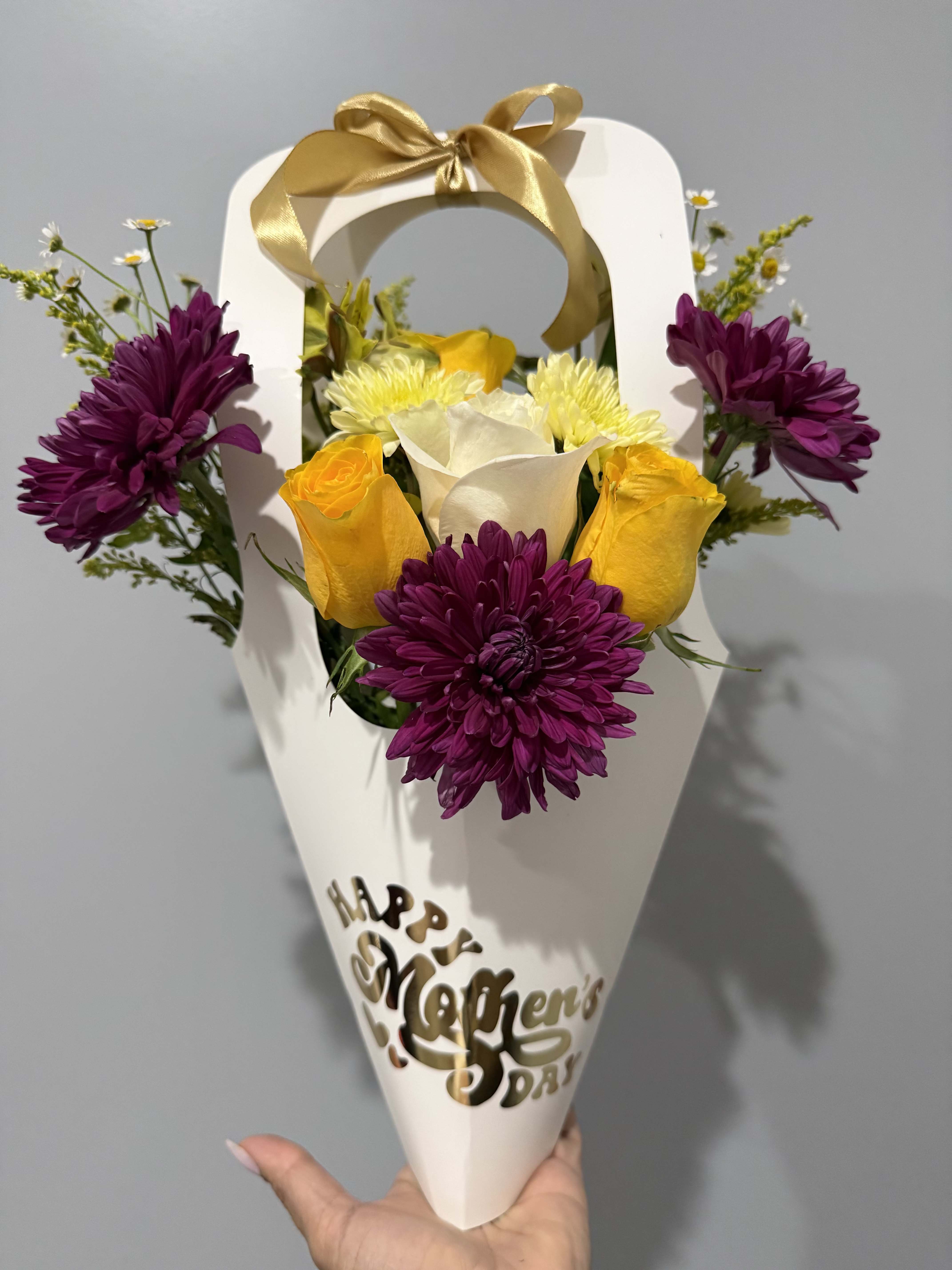 BLOOM MOM / WITHE  - A bright handheld cone of flowers that reads like a joyful hug - perfect as a Mother's Day bouquet or a springtime thank-you. This flower arrangement features yellow roses and crisp white blooms at the center, framed by deep purple chrysanthemum-like flowers and delicate daisy sprigs for a playful, high-contrast look. The visible "Happy Mother's Day" cutout makes the gifting intent clear while the color mix keeps it fresh for other occasions.  Arranged inside a white paper cone gift box and tied with a gold satin ribbon, the small, easy-to-carry size feels polished and celebratory without being fussy. The mix of roses, chrysanthemums, and daisies gives a range of textures - velvety petals, rounded pompon blooms, and airy daisy tips - that reads lively in person. Ideal for the mom you want to make feel seen, or as a sunny pick-me-up for a friend; order now for same-week gifting when available and keep stems misted and cool for the best vase life after opening the cone.