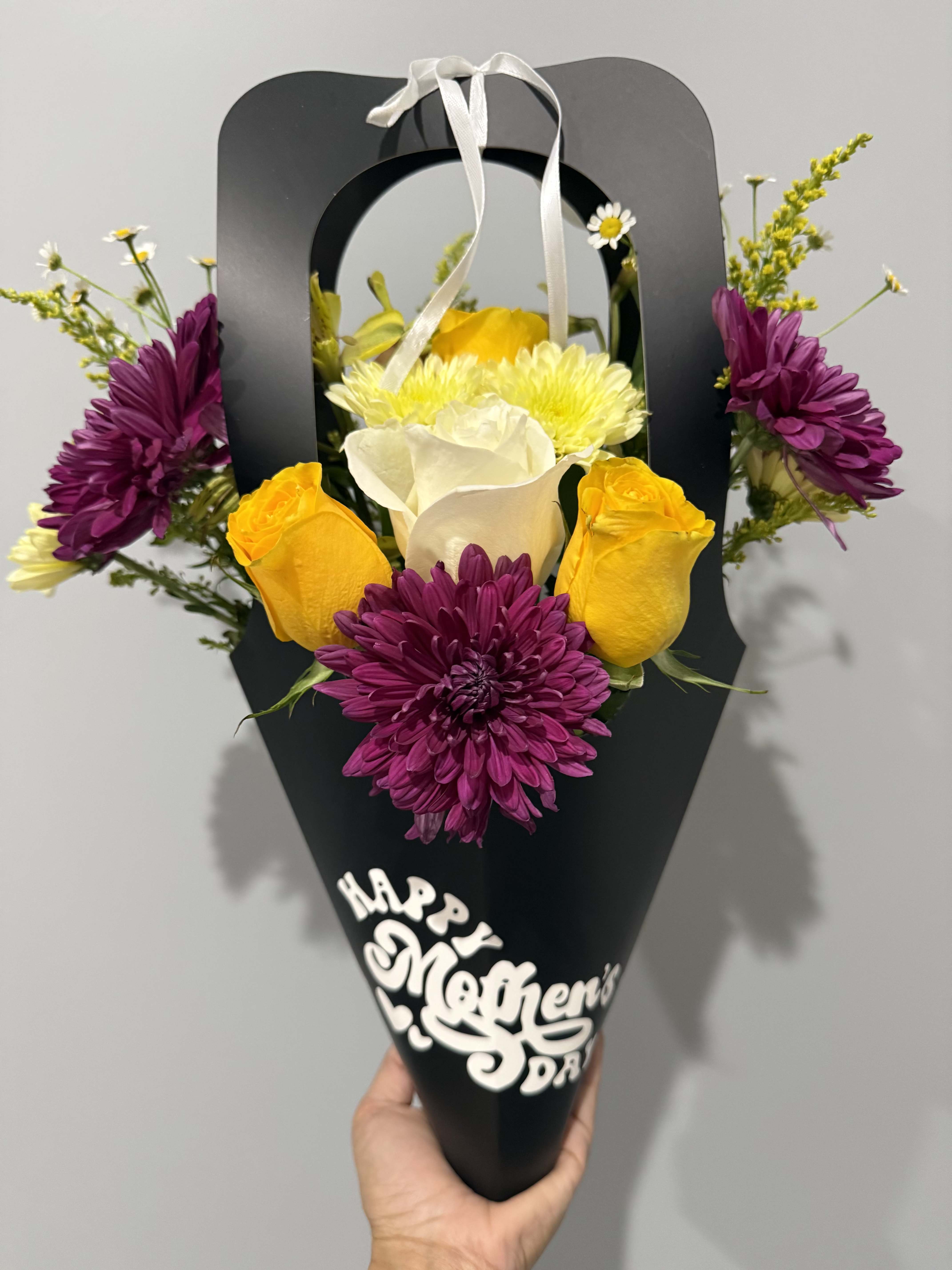 BLOOM MOM/ BLACK - Bright yellow roses and sculptural white calla lilies pop against saturated purple chrysanthemums and delicate daisy-like fillers in this hand-held flower arrangement for Mother's Day or a thoughtful appreciation bouquet. The black paper cone and white ribbon handle give the design a modern, high-contrast presentation-perfect for the mom who loves bold color and polished style. The decorative "Happy Mother's Day" cutout on the front makes the message clear without saying a word.  Carefully wrapped for gifting, the bouquet measures as a hand-held size ideal for carrying to brunch or placing in a small vase at home. Textures range from the smooth elegance of calla lilies to the ruffled charm of chrysanthemums and the classic romance of roses, creating a cheerful, celebratory feel that will make the person you're thinking of feel genuinely seen. Order ready to gift with the included black wrap and white ribbon; keep stems fresh by trimming and placing in water if transferring to a vase.
