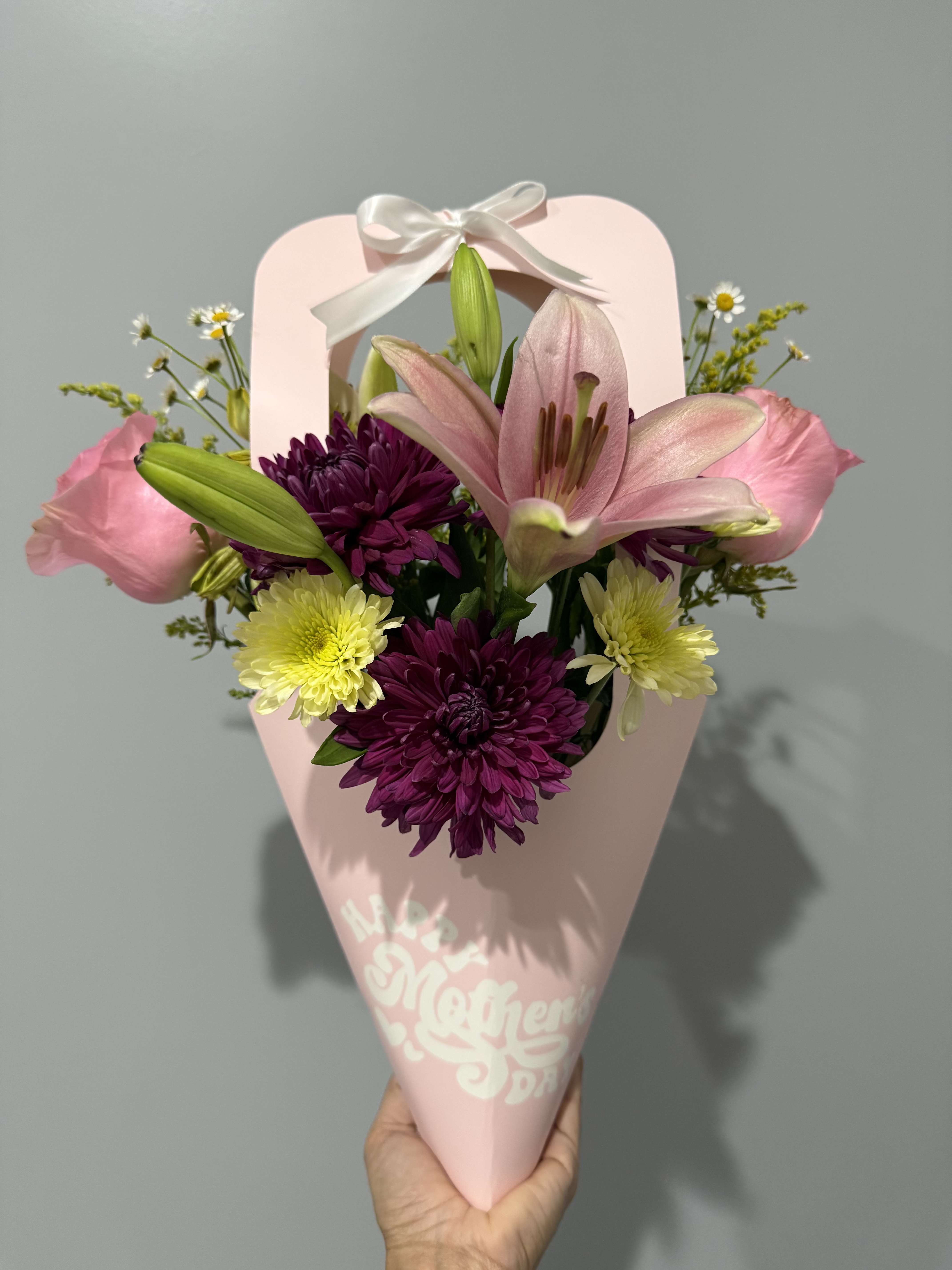 BLOOM MOM/ PINK - A big pink lily takes center stage in this cheerful hand-tied flower arrangement, making it an ideal birthday bouquet or thank-you gift that feels both feminine and fresh. The composition reads like a springtime surprise: the recipient will notice the standout lily first, then the rich purple chrysanthemums and pale yellow daisy-like blooms that brighten the mix and create joyful contrast.  Soft pink side blooms and tiny white filler daisies add lightness and a delicate texture around the bolder colors, while the compact, hand-tied shape keeps the design intimate and easy to carry. Wrapped in pastel pink paper with a white ribbon handle, the presentation arrives gift-ready and polished - the kind of bouquet that makes someone feel seen and celebrated without fuss.  Perfect for moms, friends, or anyone who loves soft, uplifting color, this arrangement suits birthdays, thank-you moments, or simply saying you're thinking of them. Size and care: compact, cone-wrapped bouquet - keep in fresh water and trim stems if you transfer to a vase to extend vase life.