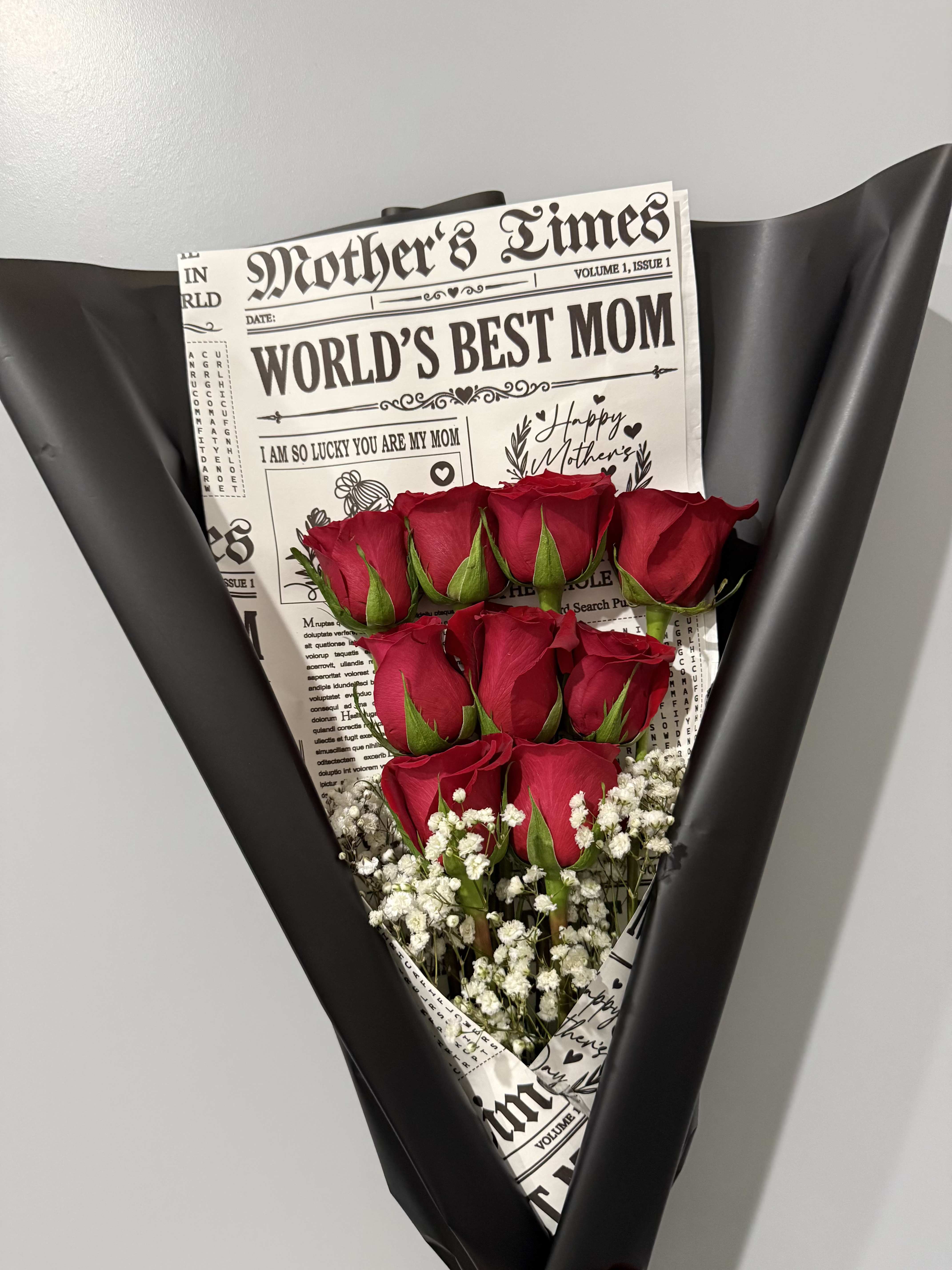 MOM BOUQUET - Deep, velvety red roses gathered in a compact, upright cluster create a dramatic, romantic Mother's Day bouquet that reads like a bold, loving statement. The arrangement is wrapped in matte black paper for a striking contrast, with a newspaper-style insert featuring "World's Best Mom" and Mother's Day wording that makes the gift feel personal and celebratory - the kind of bouquet that makes her feel truly seen and adored.  Small white filler flowers tuck around the base of the roses, adding delicate texture and bright highlights against the dark wrap. A printed newspaper-style wrap doubles as a keepsake headline while the matte black finish lends modern sophistication. Perfect for Mother's Day, an anniversary, or any moment you want to honor a mom with something both elegant and unexpectedly bold; available in a compact size ideal for delivery or hand-giving and ready to brighten her day with minimal care.