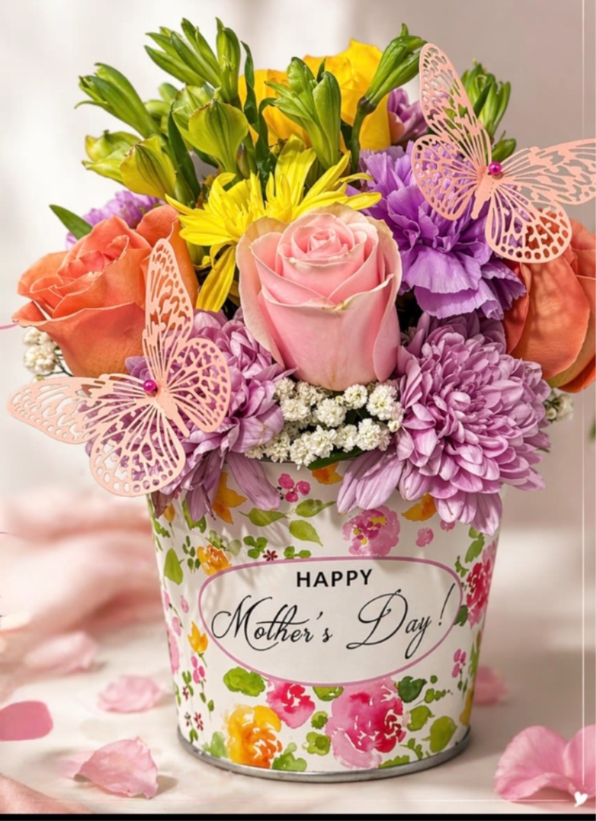 SWEET MOM - A soft pink rose takes center stage in this cheerful Mother's Day flower arrangement, surrounded by coral and orange roses, sunny yellow alstroemeria, and clusters of lavender and purple chrysanthemums. Designed as a compact, rounded bouquet for Mother's Day or a spring celebration, the bright, feminine palette and pale pink butterfly picks create a whimsical, gift-ready impression that will make the person you're thinking of feel loved and celebrated.  Flowers are tightly packed into a decorative floral-print tin with a visible "Happy Mother's Day!" message, giving the arrangement a polished, ready-to-display finish. Small white filler blooms add lightness and texture while the tin and paper butterfly picks add charming, long-lasting detail-no vase required. This arrangement arrives in a size suitable for a tabletop or bedside table and is an easy, joyful way to mark Mother's Day, a birthday, or any moment you want to make someone feel truly seen.