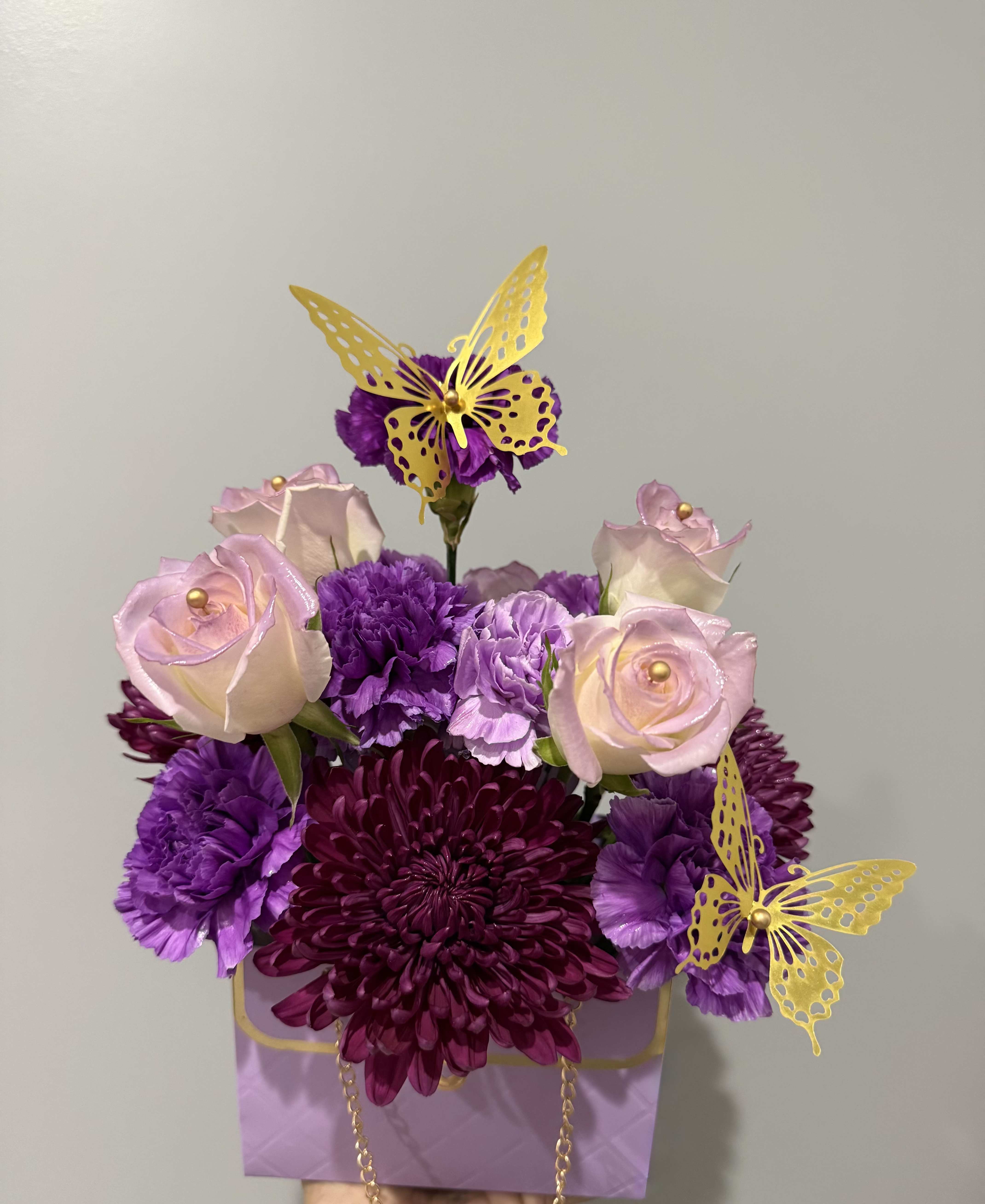 PURPLE FLOWER BAG  - Deep purple chrysanthemums and carnations mingle with pale pink roses and soft lavender blooms in a compact floral arrangement that feels both romantic and playful - perfect as a birthday bouquet, a thank-you gift, or a bright spring celebration arrangement. The purple, pink, yellow, and white palette is richly layered so the person you're thinking of will notice the depth of color at first glance and the cheerful accents up close.  The flowers are gathered in a lavender gift bag with a polished gold chain handle for easy gifting and display, while bright yellow butterfly picks rise above the blooms as whimsical focal points. Carnations and chrysanthemums provide lush texture, roses add classic romance, and the bag presentation makes this arrangement ready to set on a table or bedside without a vase.  Send this to mark a special day or simply to let someone feel seen and celebrated - it arrives ready to present, with a compact size that fits on mantels, desks, or countertops. Care: keep blooms in a cool spot out of direct sun and mist as needed to keep petals fresh.