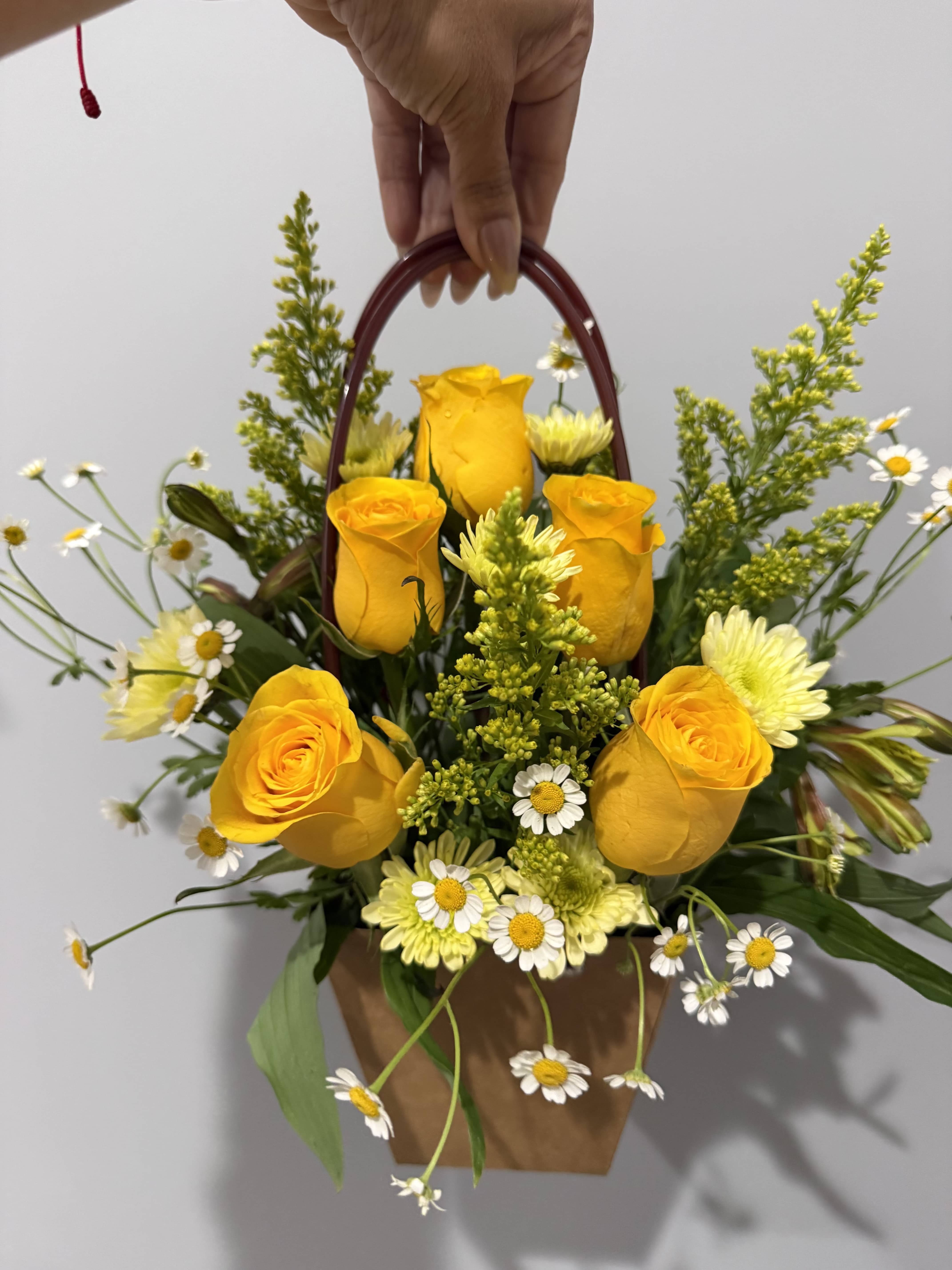 ENCANTO AMARILLO  - Sunlit yellow roses take center stage in this charming flower arrangement, a cheerful everyday bouquet perfect for birthdays, thank-you gestures, or "just because" moments. The bright yellow and white palette reads sunny and uplifting, making the person you're thinking of feel genuinely noticed and cheered the moment they see it.  Clustered with pale yellow chrysanthemums and small white daisy-like blooms, the blooms form a loose, garden-style silhouette with airy greenery and filler for a fresh, informal texture. Presented in a handled brown paper basket with an arched handle, the arrangement is easy to carry and display-an inviting mix of roses, daisies, chrysanthemums, and complimentary greens that looks effortless on a kitchen table or entryway.