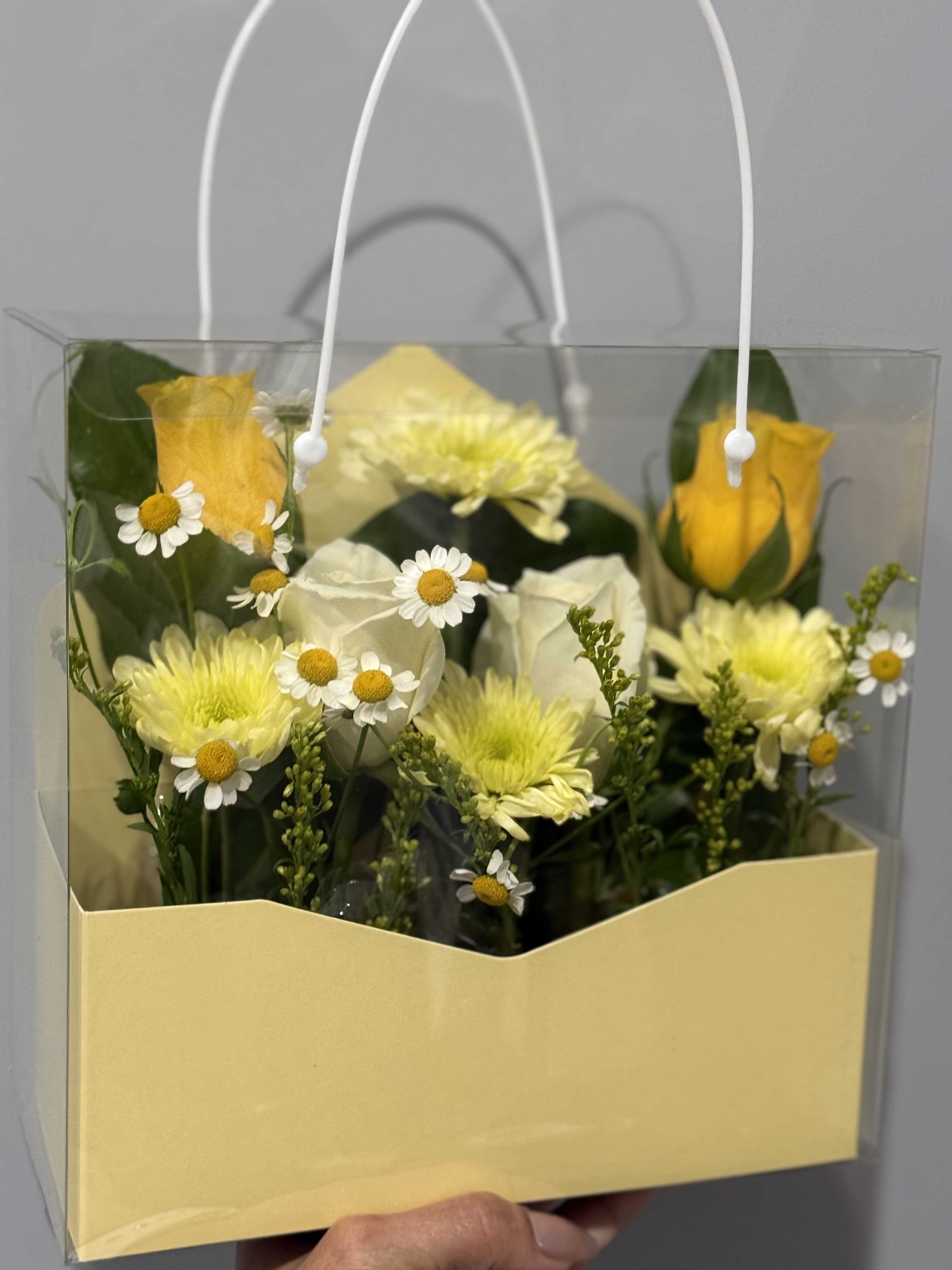 YELLOW BLISS  - Sunshine-soft yellow rose buds and pale yellow chrysanthemums meet petite white daisies in a clear handled gift bag for a spring-ready flower arrangement perfect as a birthday bouquet, thank-you gift, or everyday pick-me-up. The mix of rounded rose and chrysanthemum blooms with airy daisy accents creates a light, cheerful presence that will make the person you're thinking of feel brightened and genuinely seen.  Arranged on a pale yellow paper insert inside a clear plastic bag with white rope handles, this compact presentation is both pretty and portable-easy to hand off the doorstep or carry to a celebration. Colors are soft yellow, white, and fresh green foliage; the variety of textures gives the bouquet depth without feeling fussy. Order now for a thoughtful spring celebration or simple gesture of appreciation; this arrangement arrives ready to display as shown in the clear bag.